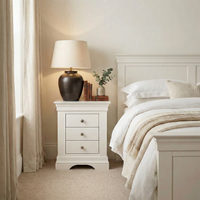 The Chalbury 3 Drawer Bedside Table in Warm White sits beside a neatly made bed, styled with a lamp, books, and a small plant to add charm and function to your neutral-toned bedroom décor.