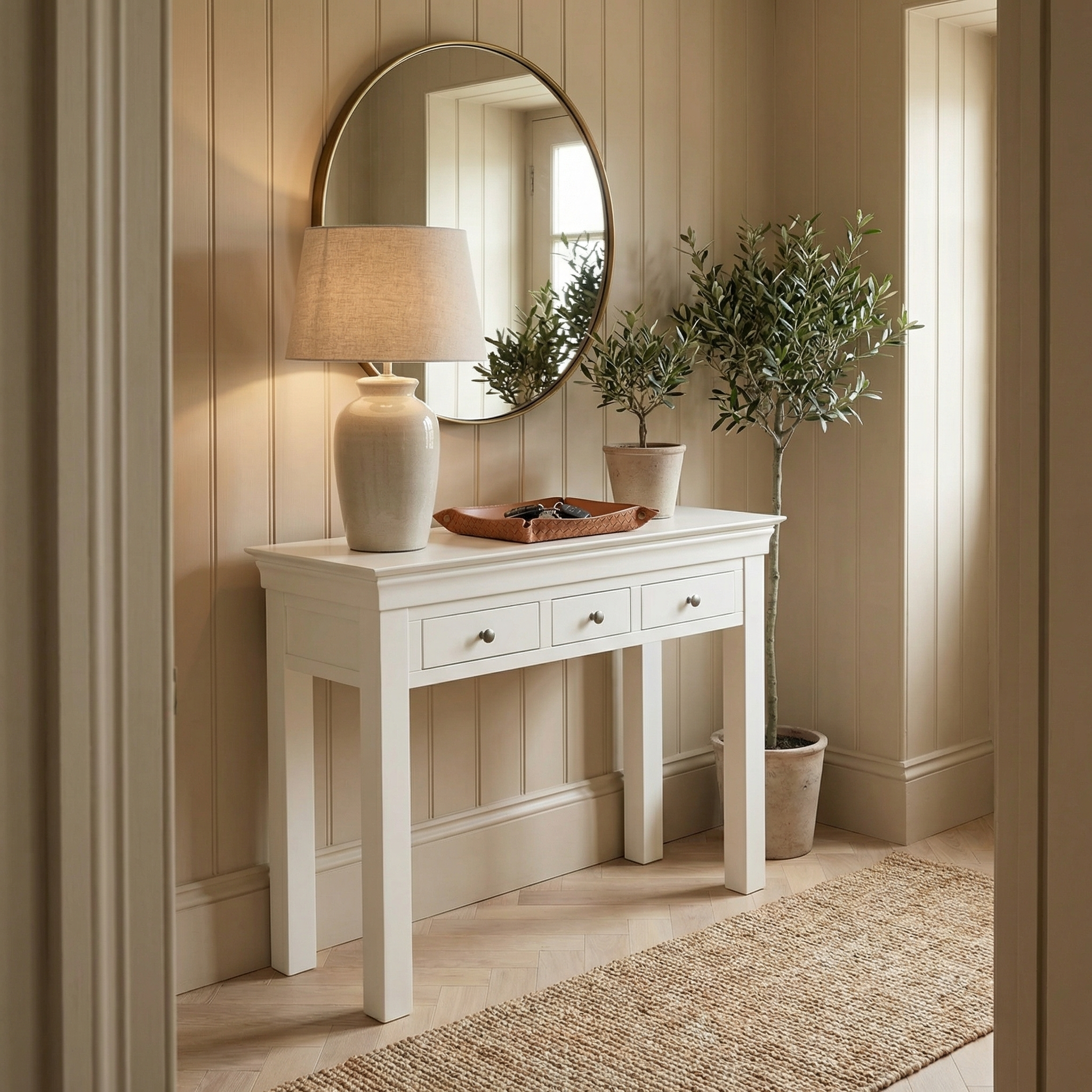 The Chalbury Console Desk in Warm White is styled with a lamp, bowl, mirror, and potted plant in a bright hallway featuring neutral decor.