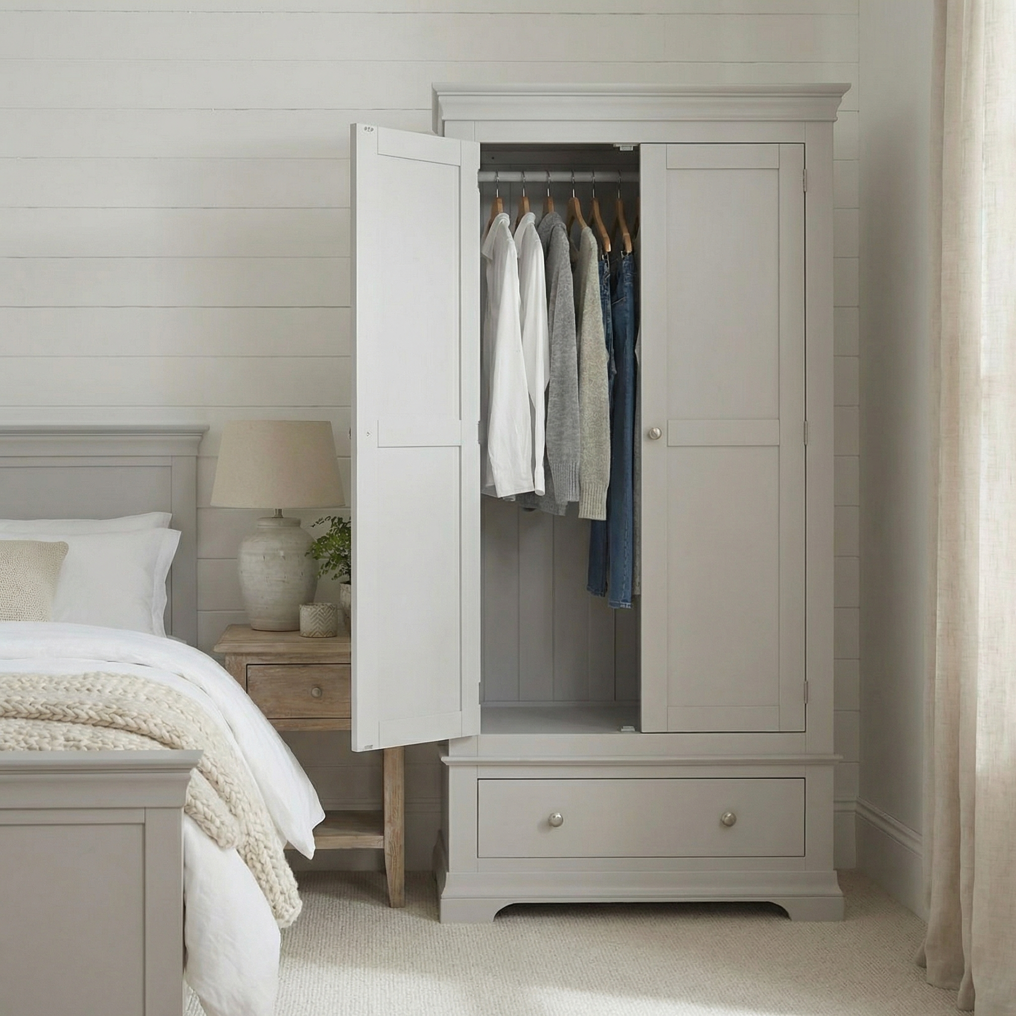 The Chalbury Double Wardrobe in Pebble Grey stands open to display hanging clothes, providing practical bedroom storage next to a bed and nightstand.
