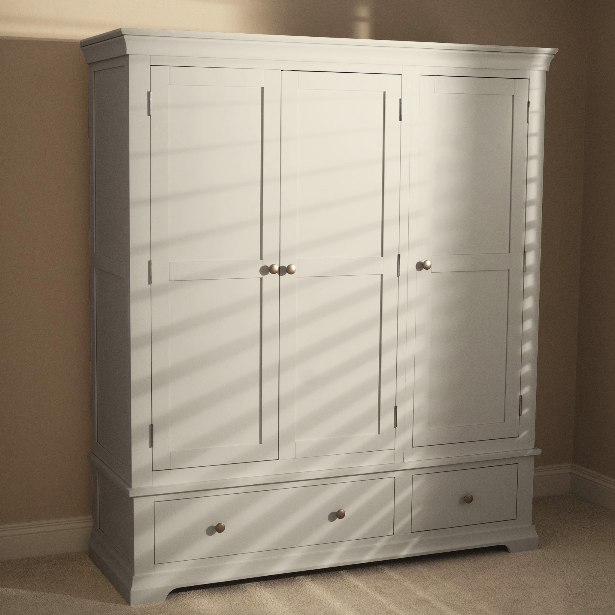 Sunlight casts striped shadows across the Chalbury Triple Wardrobe in Pebble Grey, a wooden bedroom wardrobe featuring three doors and two drawers.