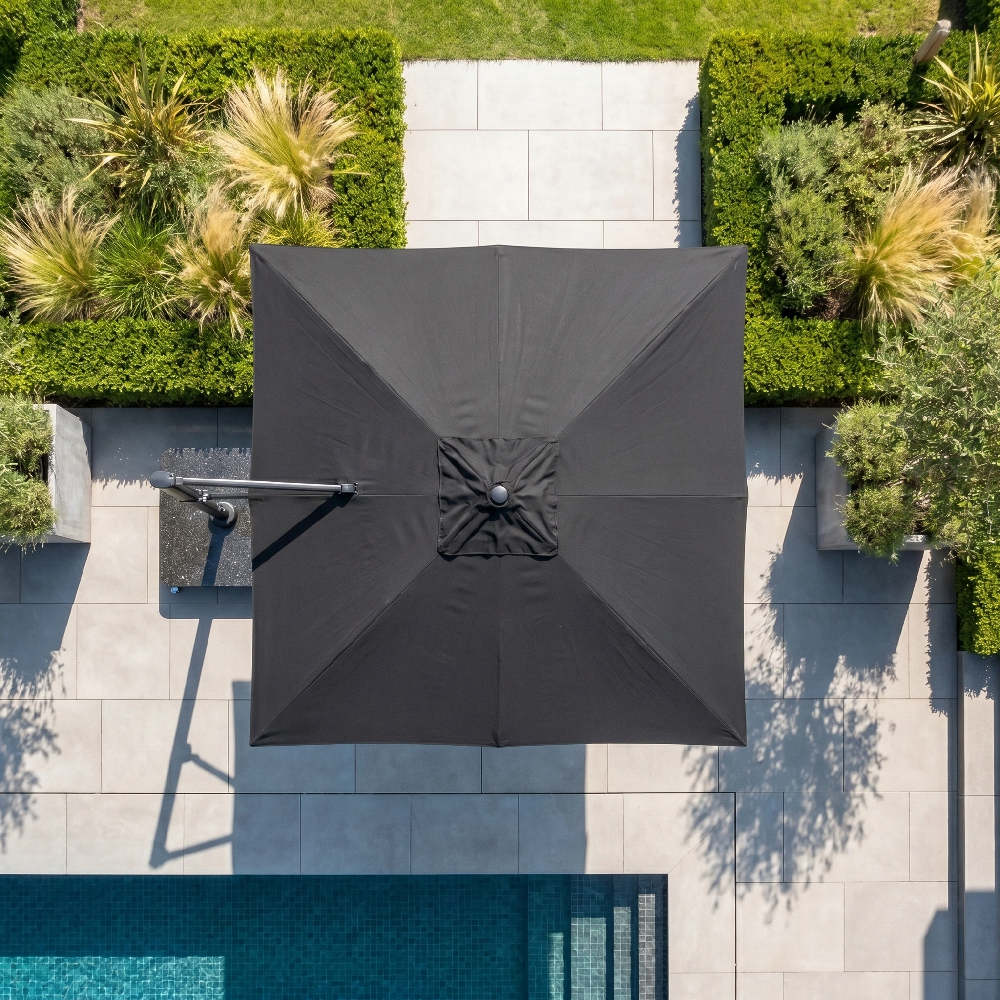 The Challenger T2 Premium 3.5m x 2.6m Rectangular Cantilever Parasol in Black shades a poolside area with tiled paving and green hedges, seen from above.