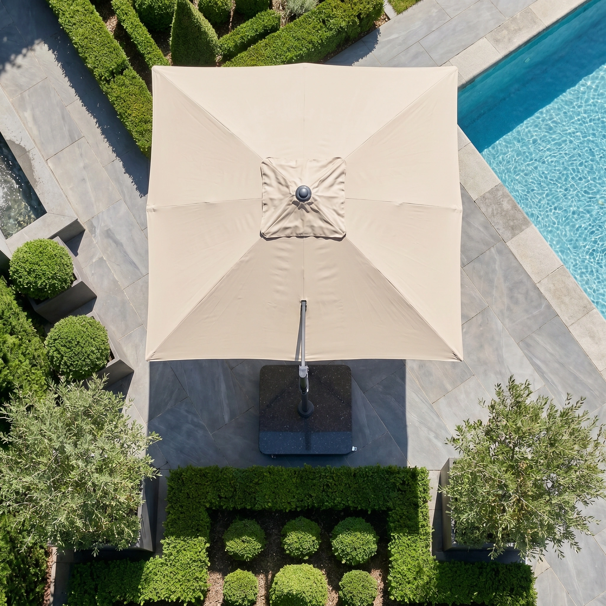 The Challenger T2 Premium 3.5m x 2.6m Rectangular Cantilever Parasol in Champagne offers stylish outdoor shade, viewed from above among lush greenery beside a blue swimming pool.