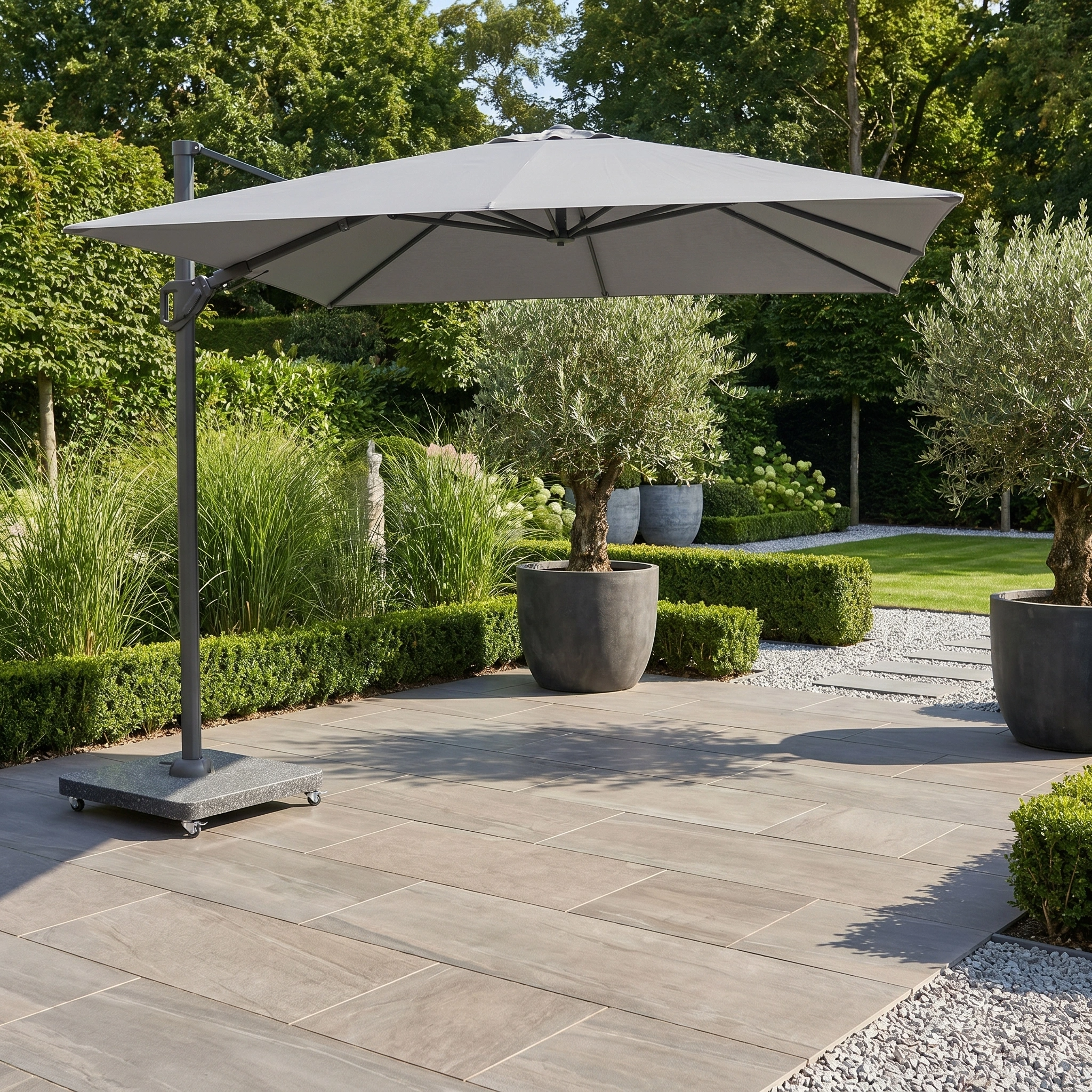 The Challenger T2 Premium 3.5m x 2.6m Rectangular Cantilever Parasol in Manhattan Grey shades a tiled outdoor area with potted trees and a green garden in the background.