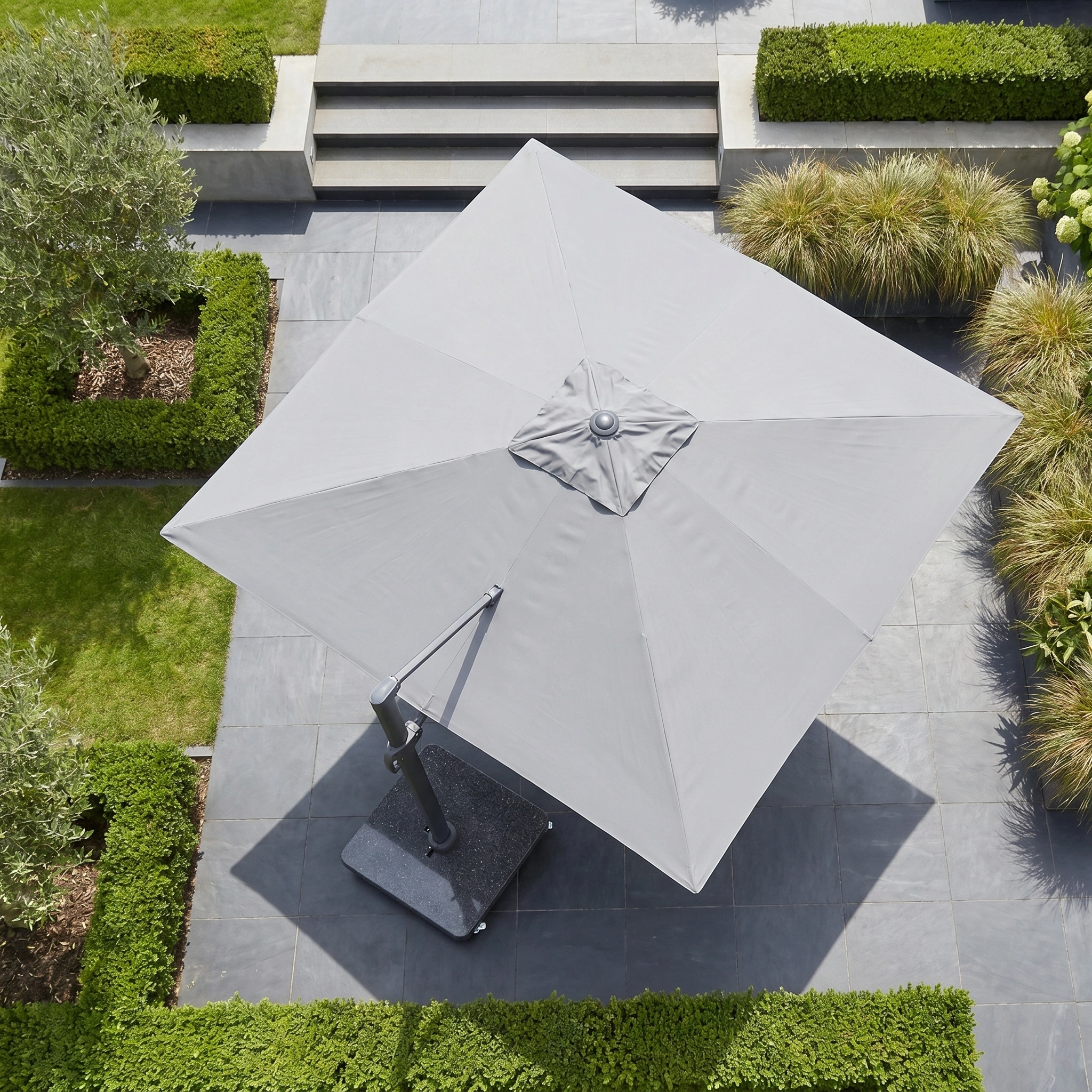 The Challenger T2 Premium 3.5m x 2.6m Rectangular Cantilever Parasol in Manhattan Grey stands on a tiled terrace, providing outdoor shade among trimmed hedges and lush plants.