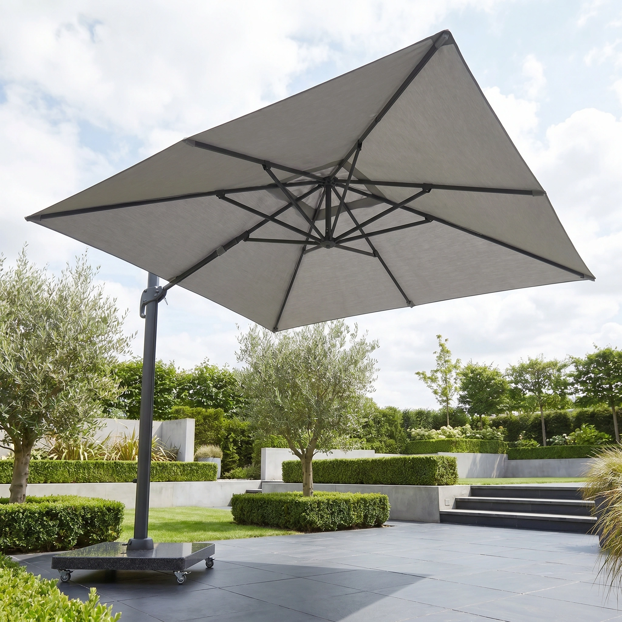 The Challenger T2 Premium 3.5m x 2.6m Rectangular Cantilever Parasol in Manhattan Grey provides outdoor shade on a tiled terrace, set within a landscaped garden featuring bushes and trees.