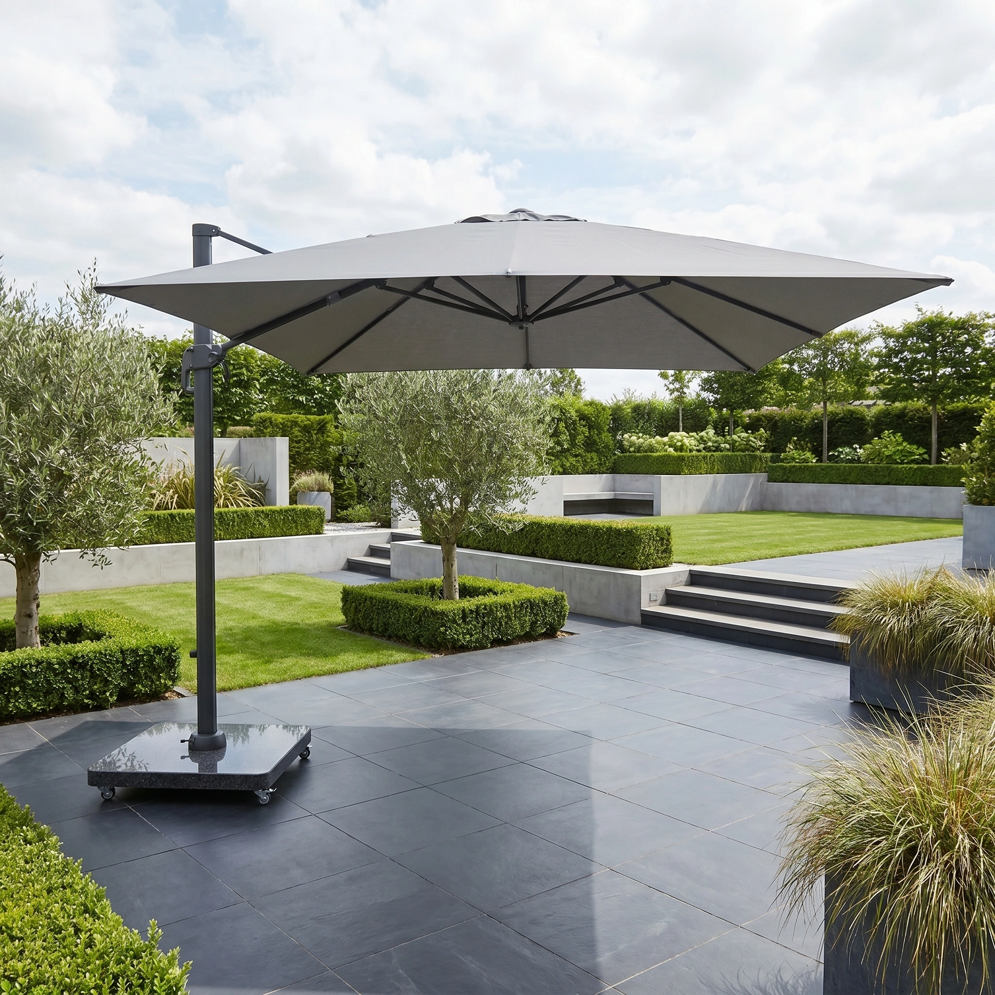 The Challenger T2 Premium 3.5m x 2.6m Rectangular Cantilever Parasol in Manhattan Grey stands on a tiled terrace in a modern landscaped garden with hedges and trees, providing stylish outdoor shade.