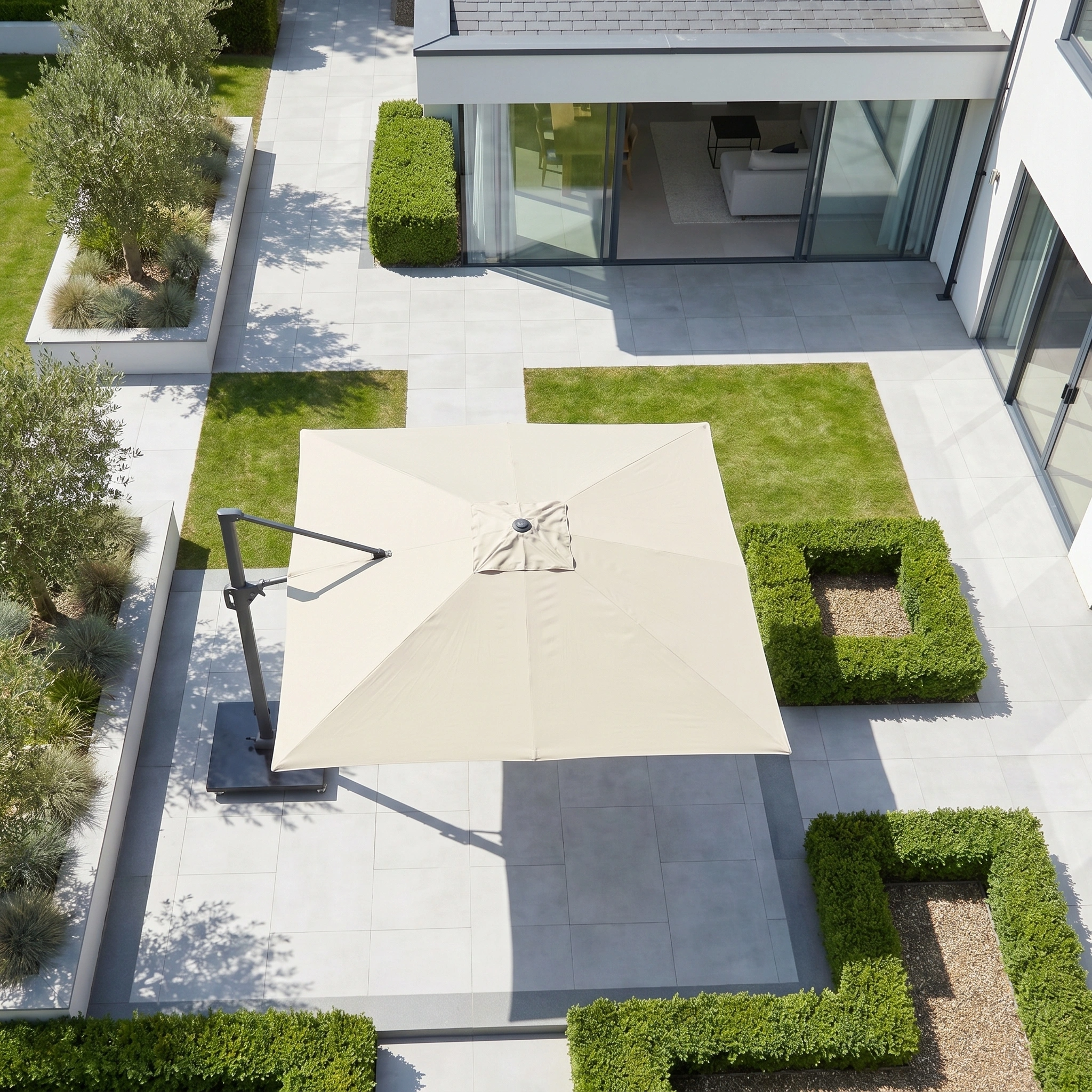 A modern patio features the Challenger T2 3.5m x 2.6m Rectangular Cantilever Parasol in Champagne, green hedges, and tiled flooring beside a white building for stylish outdoor shade.