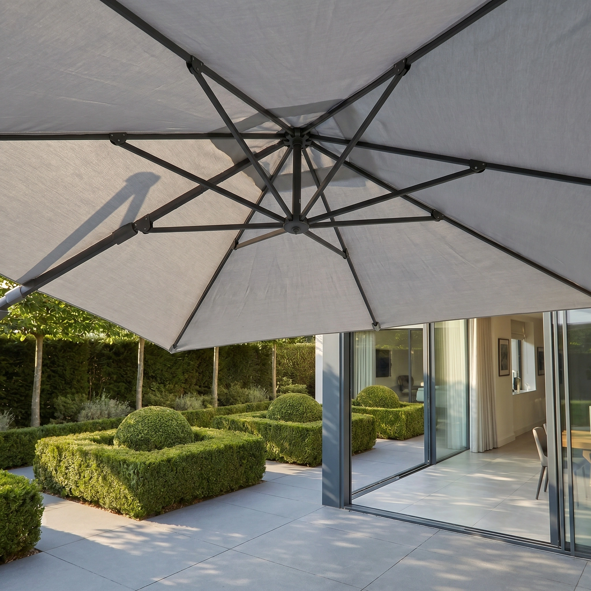 The Challenger T2 3.5m x 2.6m Rectangular Cantilever Parasol in Grey, featuring a robust 90kg granite base, stands elegantly by the garden's neat hedges and sliding glass doors.