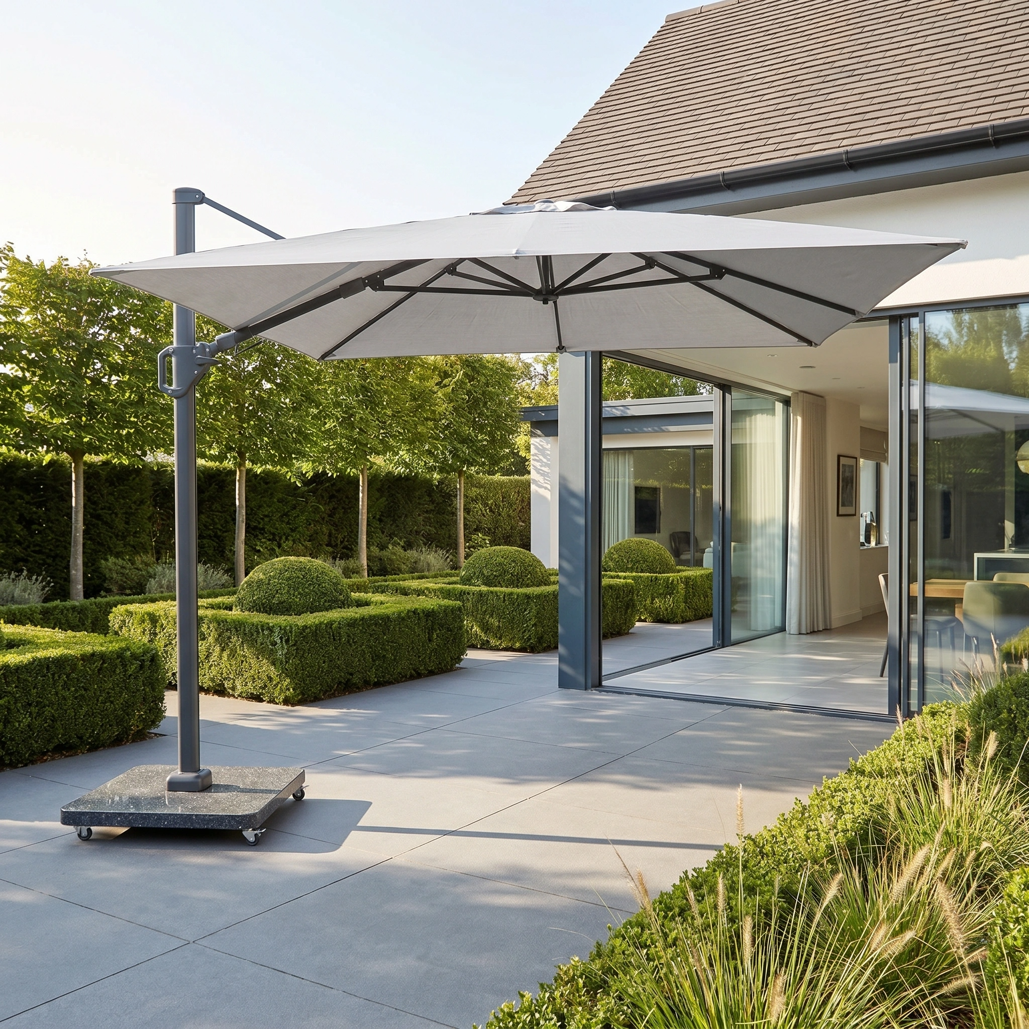 The Challenger T2 3.5m x 2.6m Rectangular Cantilever Parasol in Grey with a 90kg granite base stands elegantly over a modern patio and manicured garden.