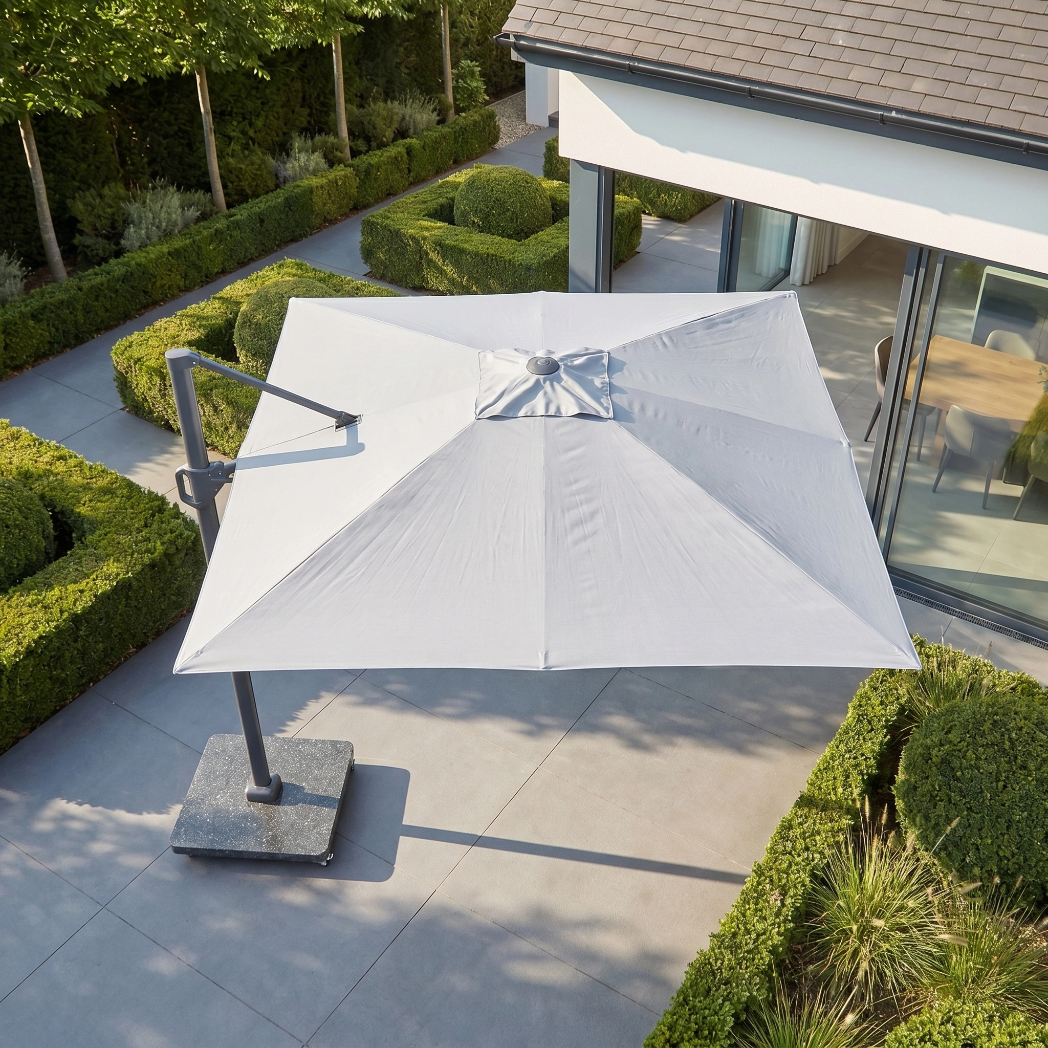 The Challenger T2 3.5m x 2.6m Rectangular Cantilever Parasol in Grey with a 90kg granite base shades a modern outdoor area with greenery and glass doors.