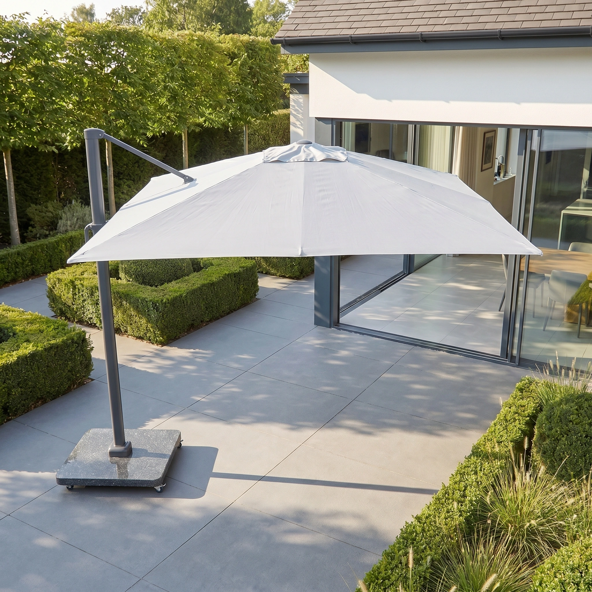 The Challenger T2 3.5m x 2.6m Rectangular Cantilever Parasol in Grey with a 90kg granite base shades a stylish outdoor terrace filled with plants and glass doors leading into the house.