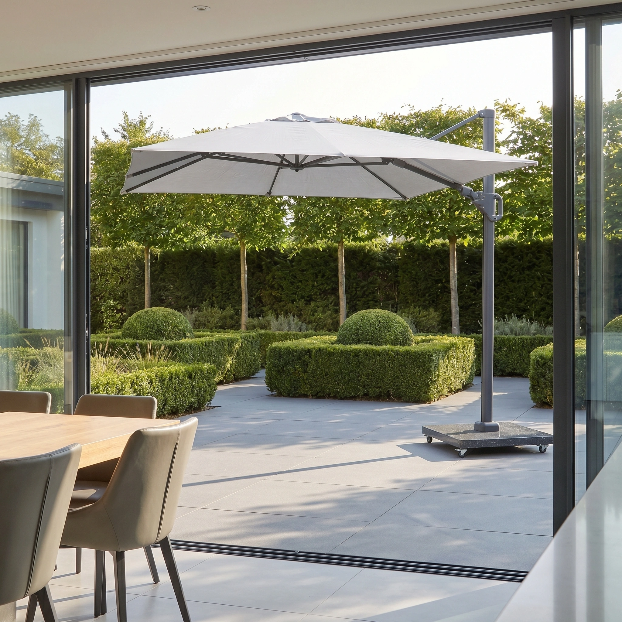 Modern patio with a Challenger T2 3.5m x 2.6m Rectangular Cantilever Parasol in Grey and 90kg Granite Base, neat hedges, and a dining set, seen through open glass doors.