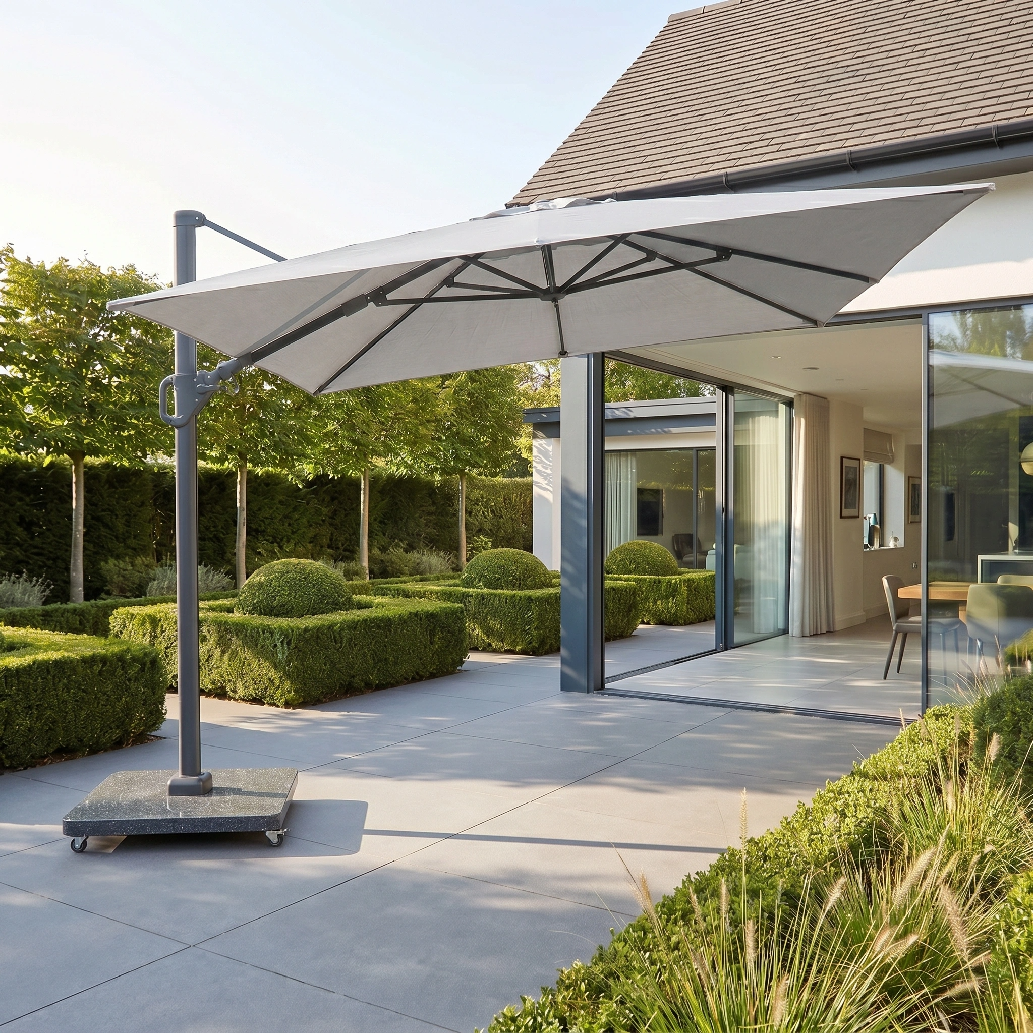 Modern patio with manicured hedges and sliding glass doors, featuring a Challenger T2 3.5m x 2.6m rectangular cantilever parasol in grey with a 90kg granite base.