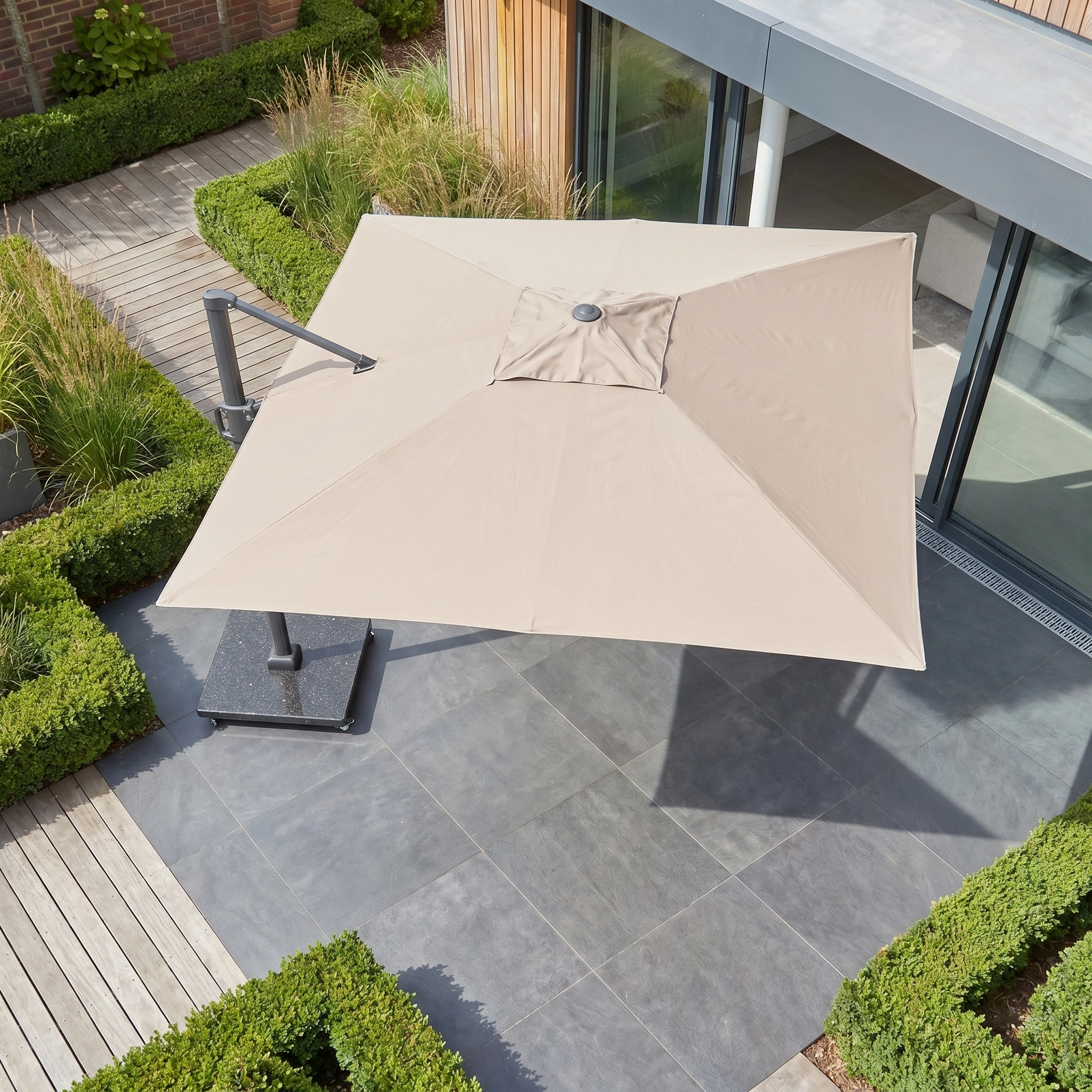 The Challenger T2 3.5m x 2.6m Rectangular Cantilever Parasol in Taupe with a 90kg granite base provides shade for a tiled outdoor area beside green hedges and a modern building.