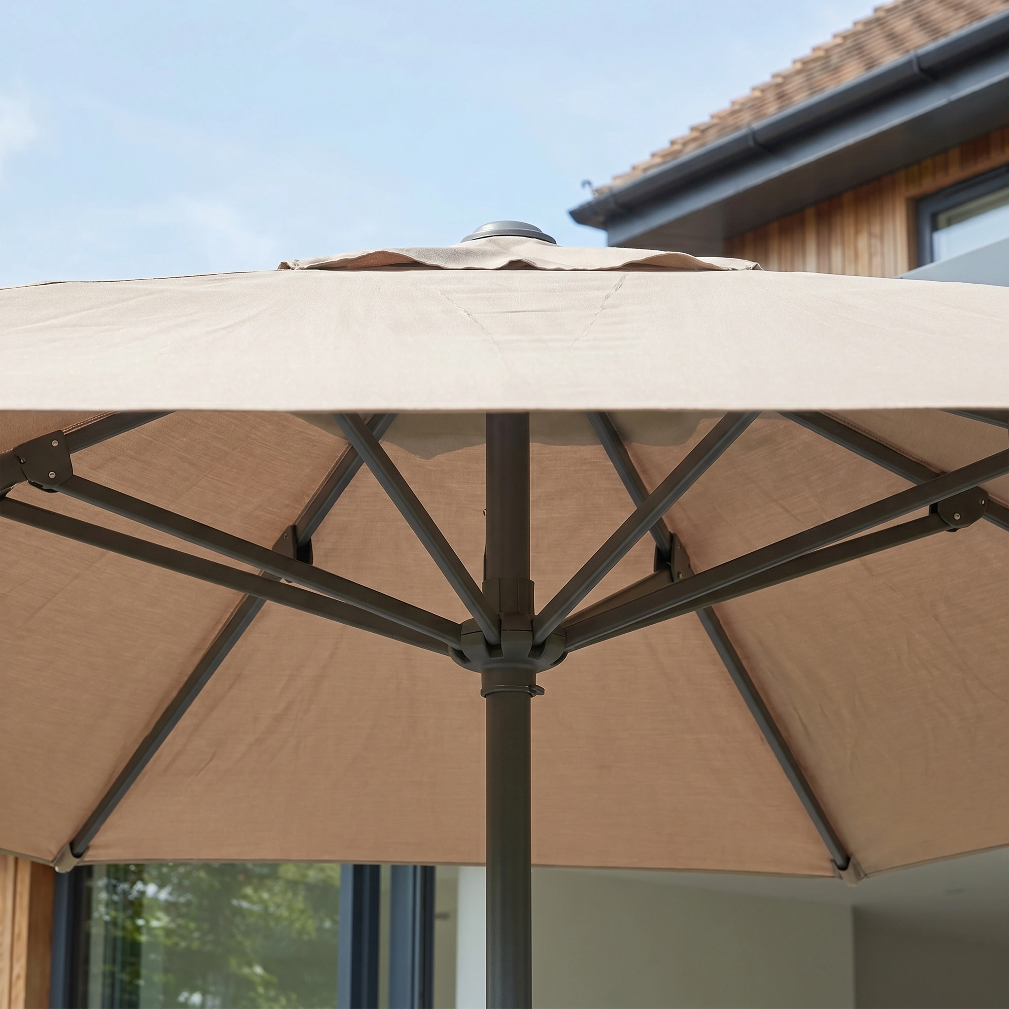 The Challenger T2 3.5m x 2.6m Rectangular Cantilever Parasol in Taupe with 90kg Granite Base is shown from below against a backdrop of a modern house and blue sky.