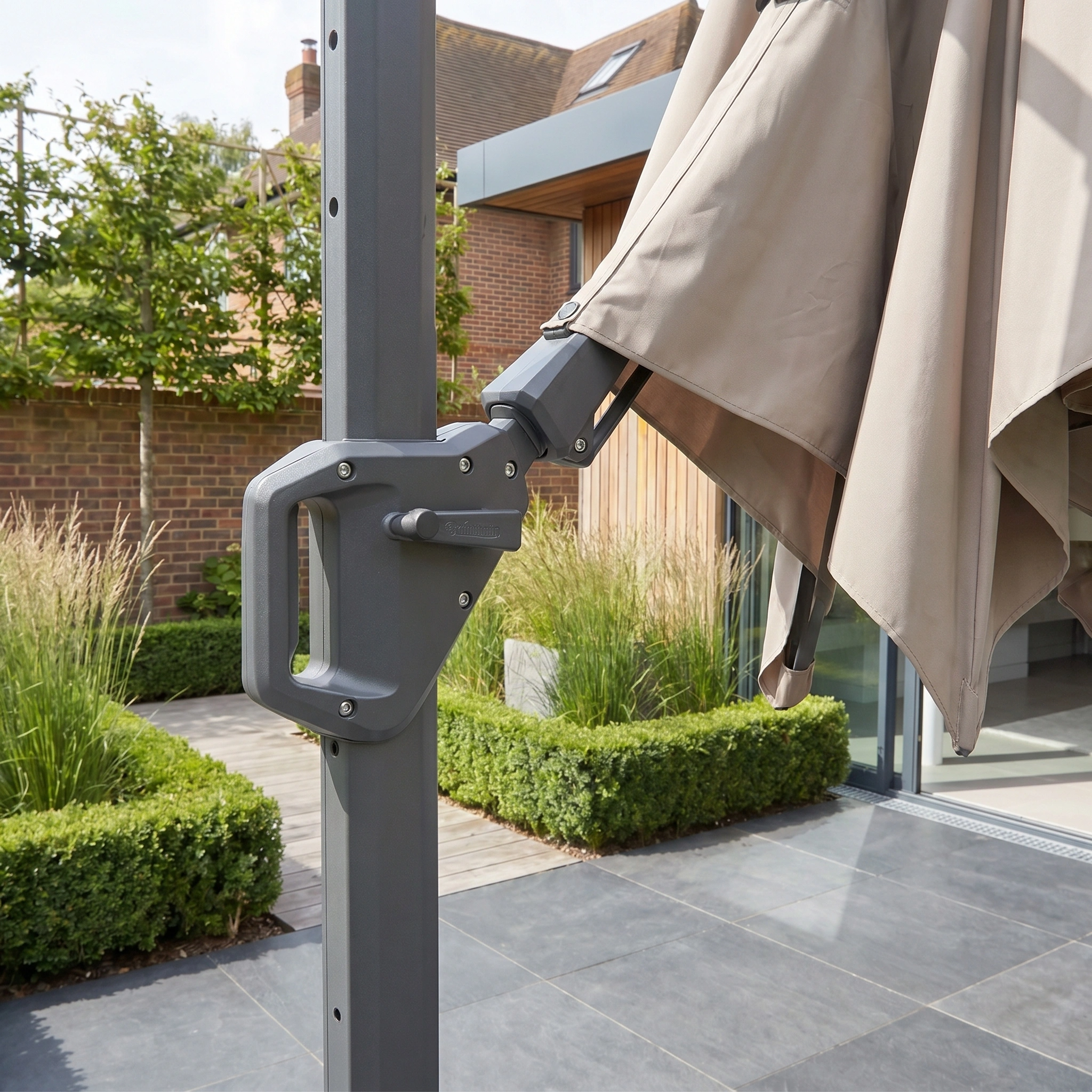 Close-up of the Challenger T2 3.5m x 2.6m Rectangular Cantilever Parasol crank handle in Taupe, shown with greenery and modern patio tiles, paired with a sturdy 90kg granite base.