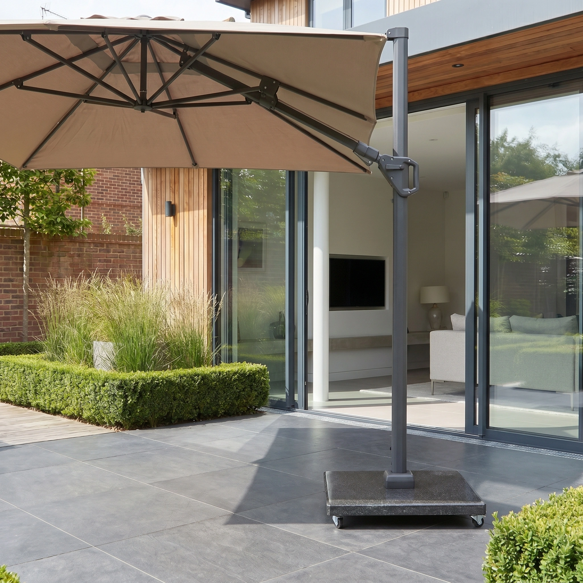 The Challenger T2 3.5m x 2.6m Rectangular Cantilever Parasol in Taupe with a 90kg granite base provides premium UV protection for your outdoor area next to sleek glass doors and neatly trimmed bushes.