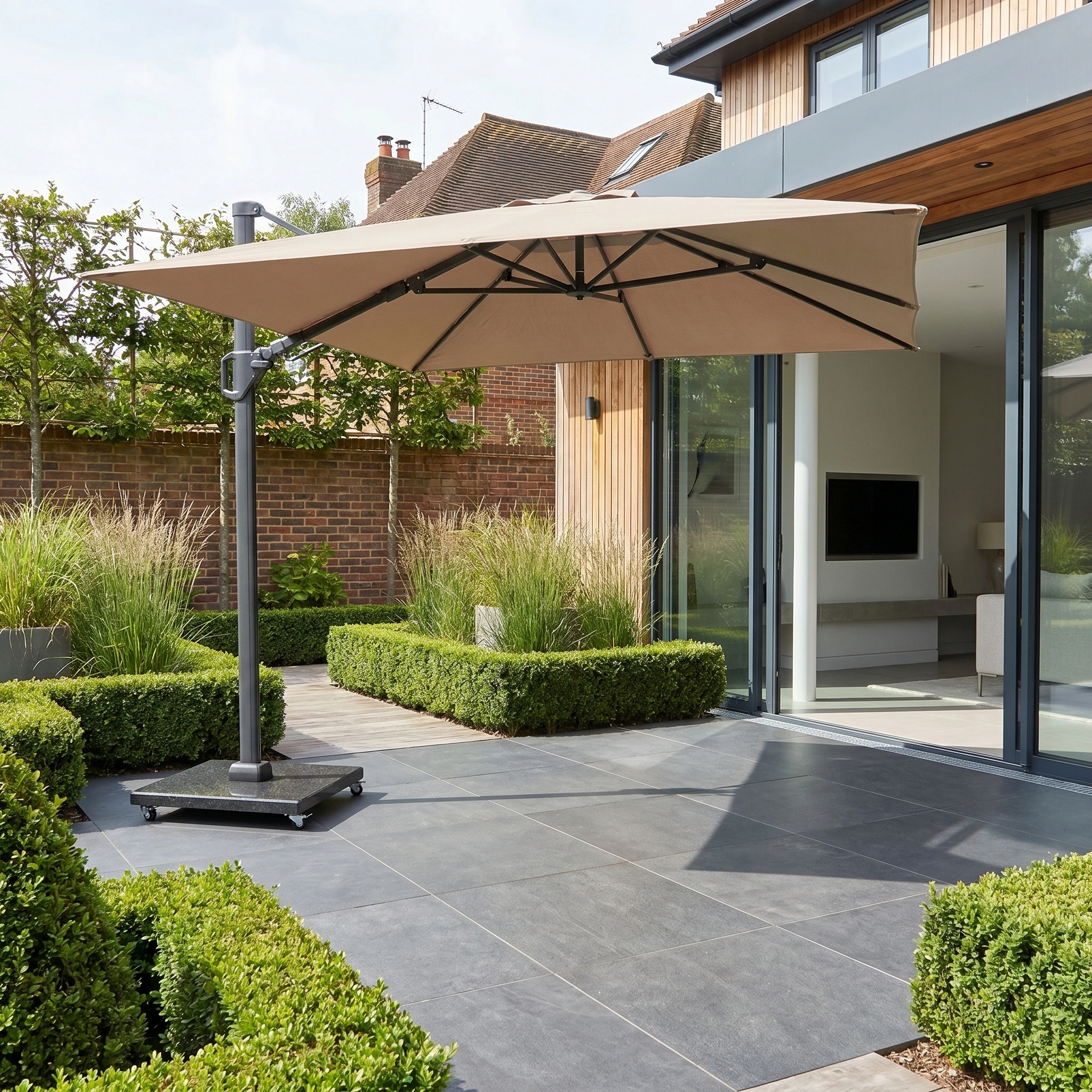 Modern patio with gray tiles, trimmed green bushes, and a Challenger T2 3.5m x 2.6m Rectangular Cantilever Parasol in Taupe with 90kg Granite Base outside a contemporary house.