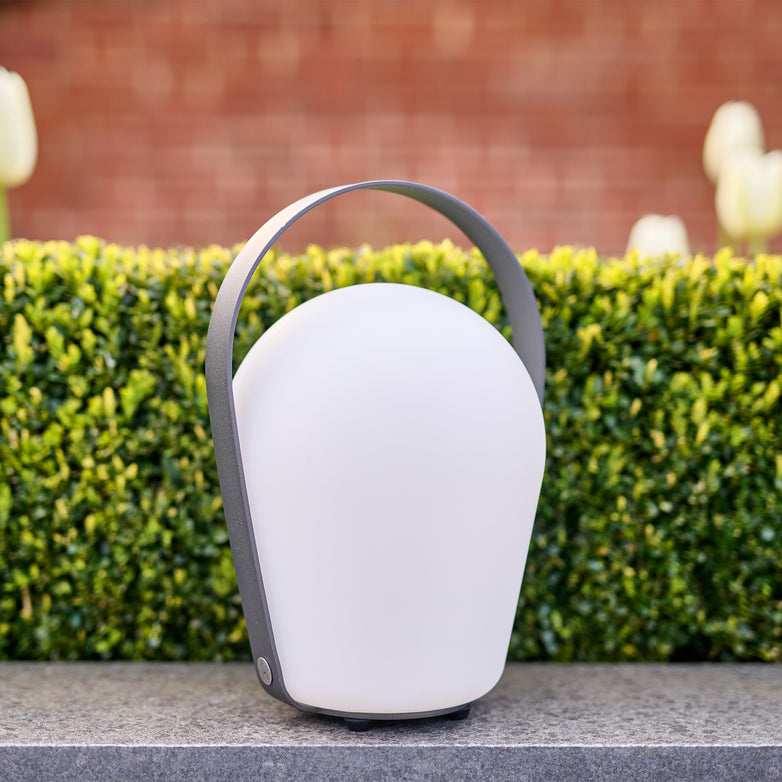 Charcoal lantern with a curved handle, placed on a stone surface in front of a green hedge and blurred background. This large Bob table light is weather resistant, making it perfect for both indoor and outdoor settings.