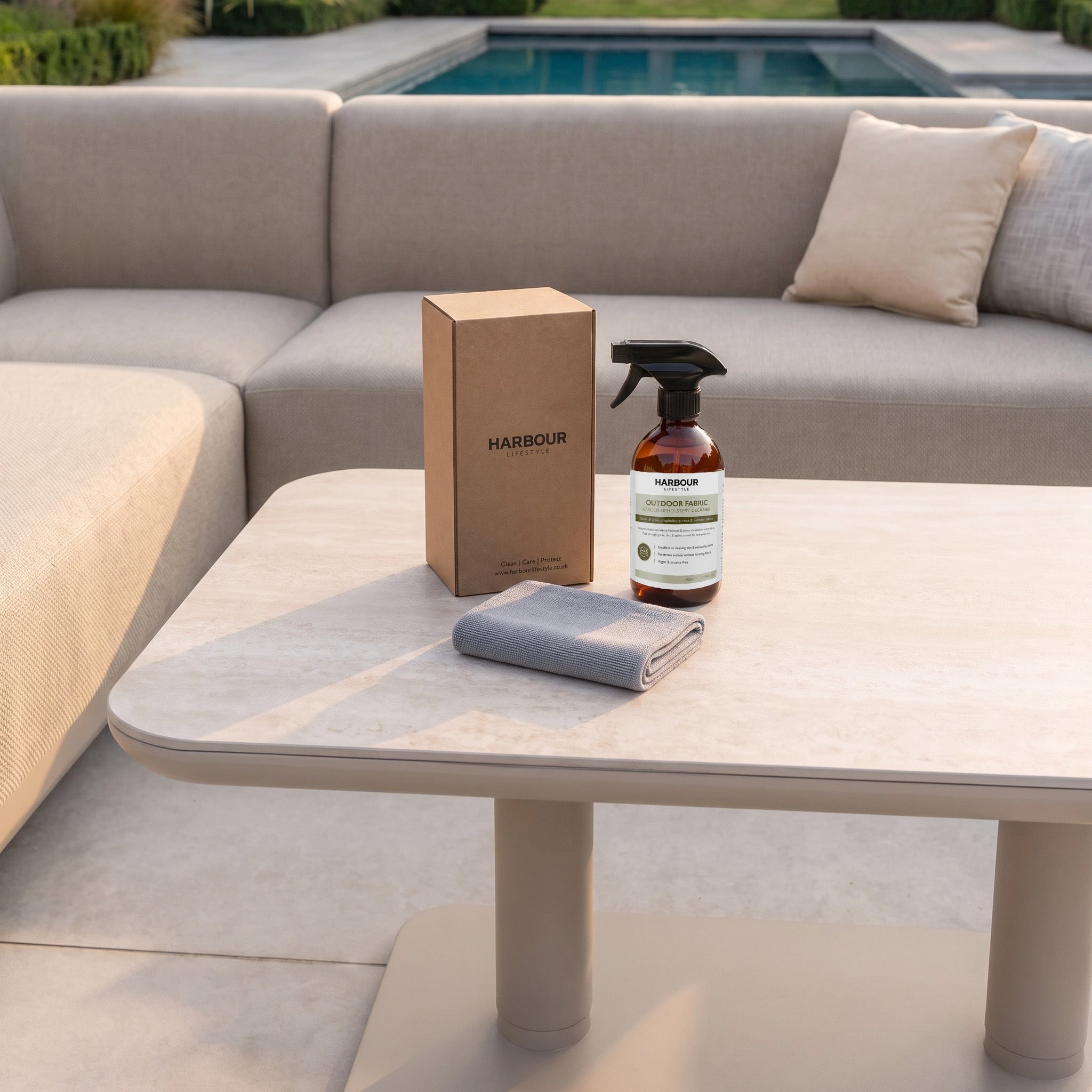 A spray bottle of Outdoor Furniture Cushion, Rope and Upholstery Cleaner, a box, and a cloth sit on a beige outdoor table by a poolside sofa set.