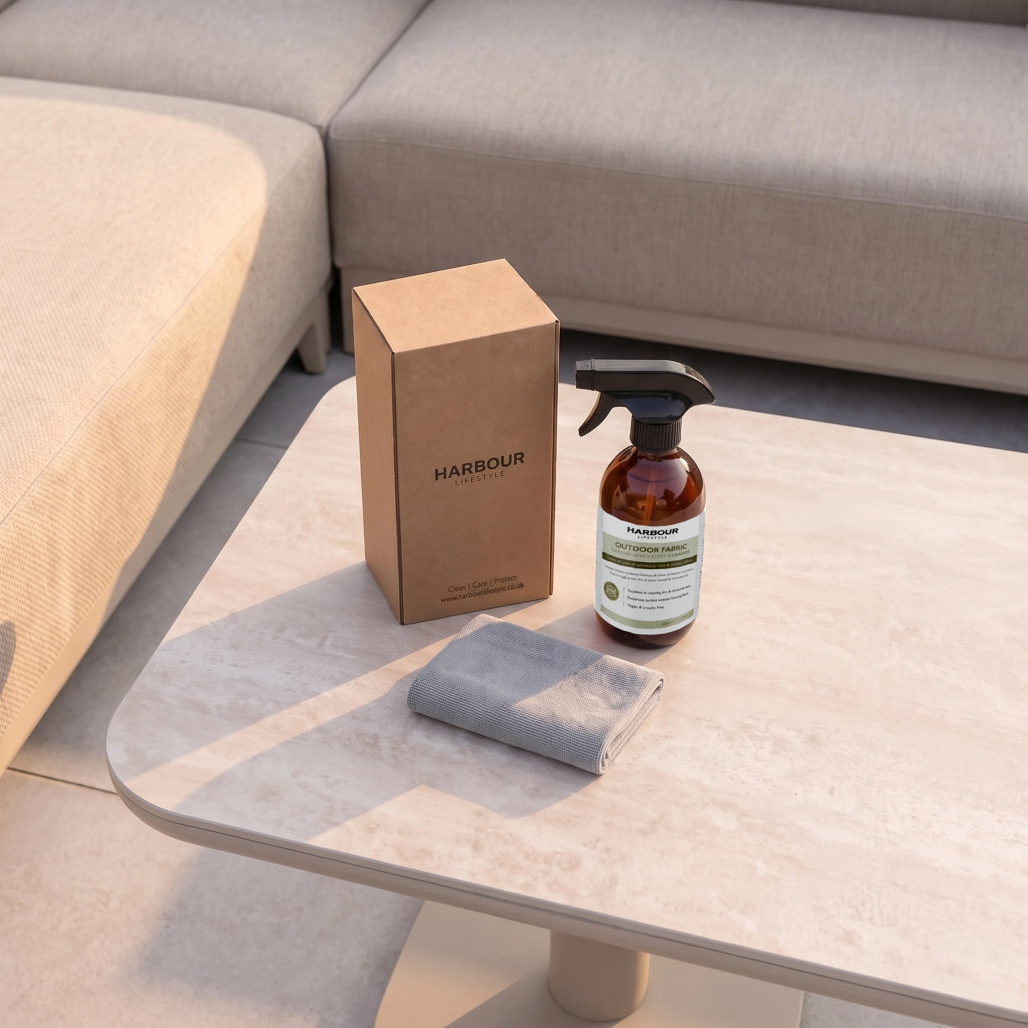 A spray bottle of Outdoor Furniture Cushion, Rope and Upholstery Cleaner, a brown box, and a gray cloth are on a light-colored table near beige couches.