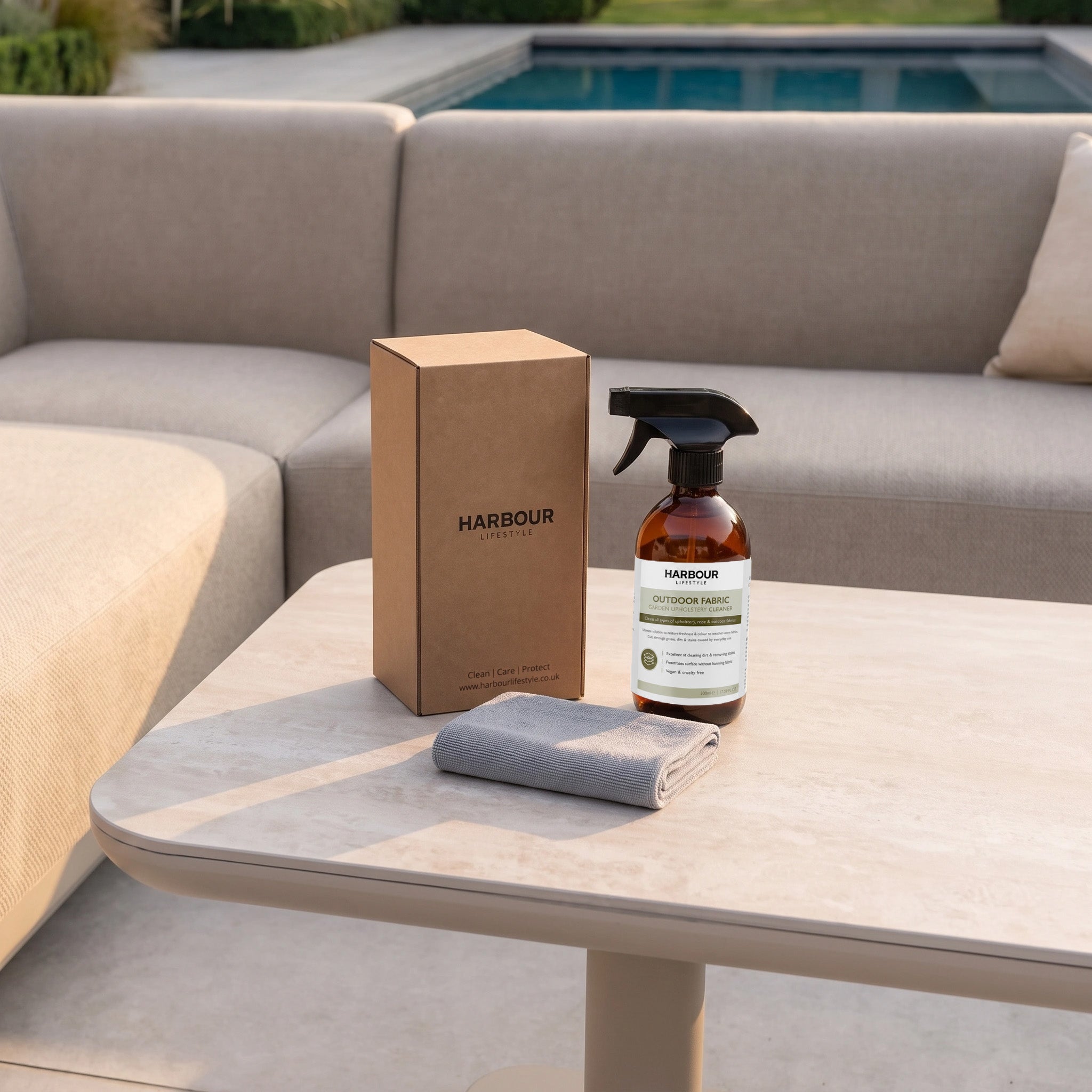 A spray bottle of Outdoor Furniture Cushion, Rope and Upholstery Cleaner, a cloth, and a box labeled “Harbour” rest on a table by an outdoor sofa and swimming pool.