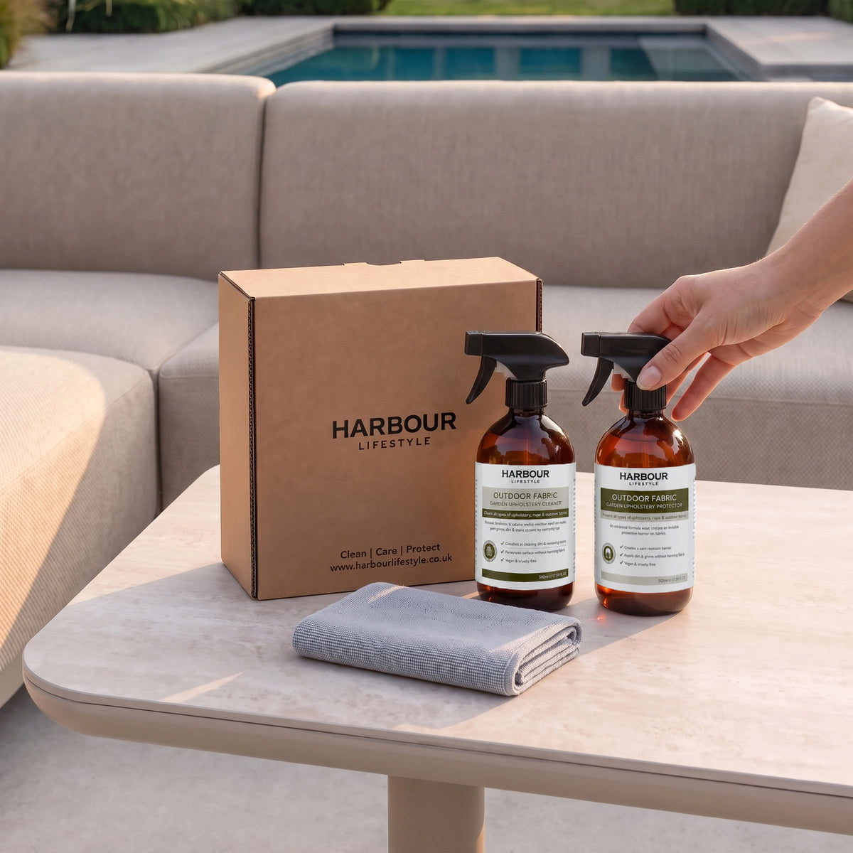 A hand places the Outdoor Furniture Cushion, Rope and Upholstery Cleaner & Protector Care Kit—complete with two Harbour Lifestyle cleaners, a box, and cloth—on a poolside table beside a sofa: your go-to essentials for cleaning garden upholstery and preventing mould and mildew.