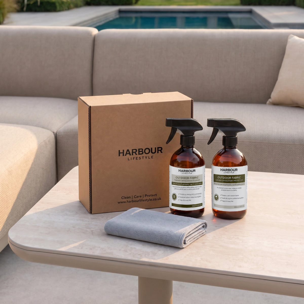The Outdoor Furniture Cushion, Rope and Upholstery Cleaner & Protector Care Kit—with two spray bottles, a box, and a folded cloth—sits on a beige table by a sofa next to the swimming pool, standing ready for effective mould and mildew prevention.