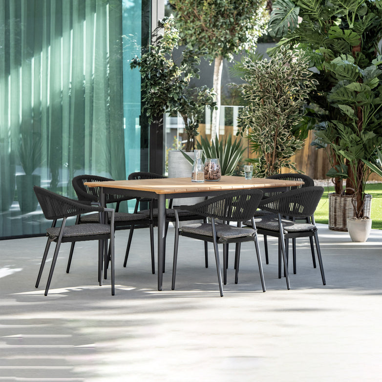 The Cloverly 6 Seat Rectangular Dining with Teak Table in Charcoal offers a stylish outdoor dining experience, featuring a teak top wooden table and sleek black chairs with aluminium frames, all elegantly positioned on a patio surrounded by lush green plants.