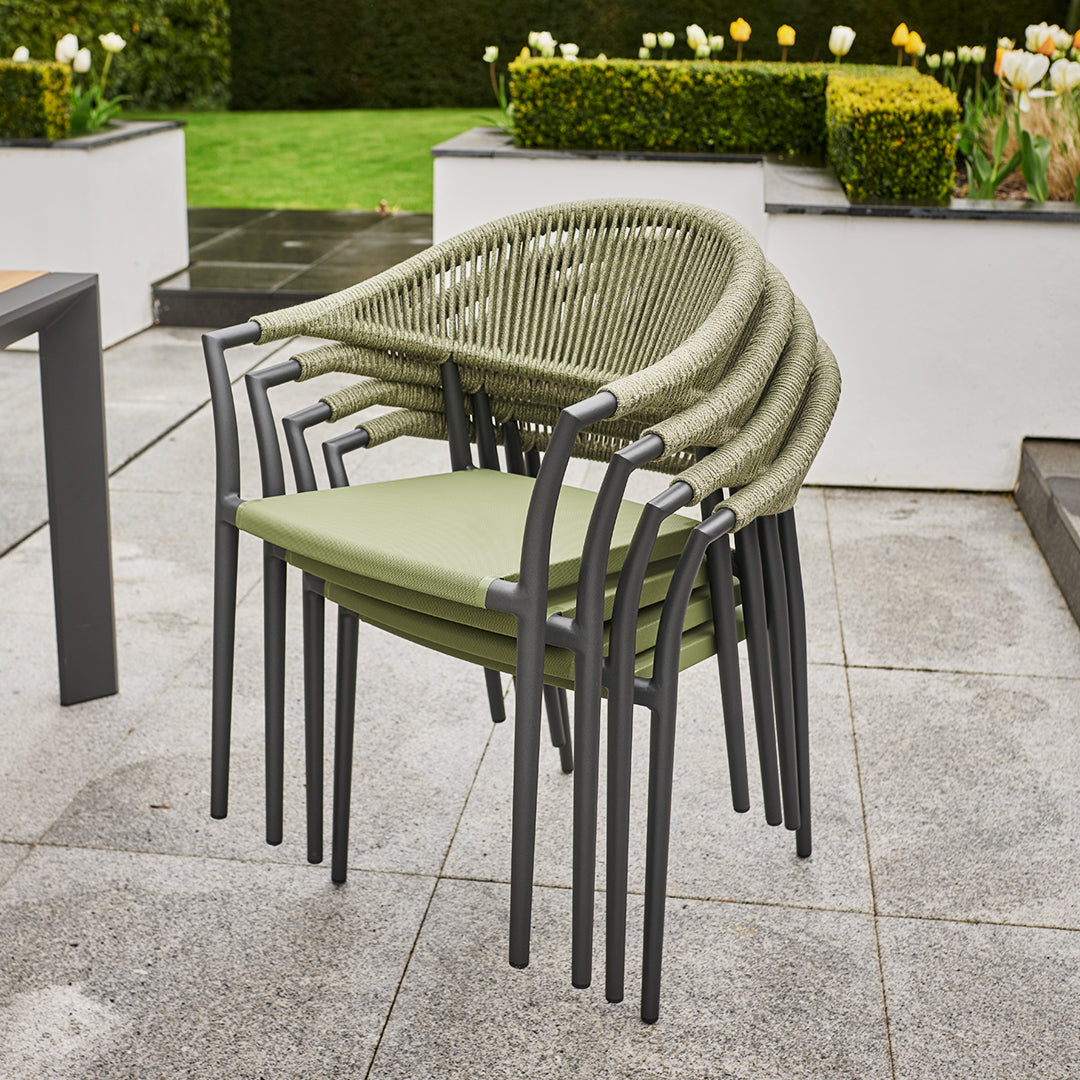 Four stacked Cloverly outdoor chairs with olive green seats and rope backs stand beside the ceramic glass table from the 8 Seat Rectangular Extending Dining Set, framed by garden plants on the patio.