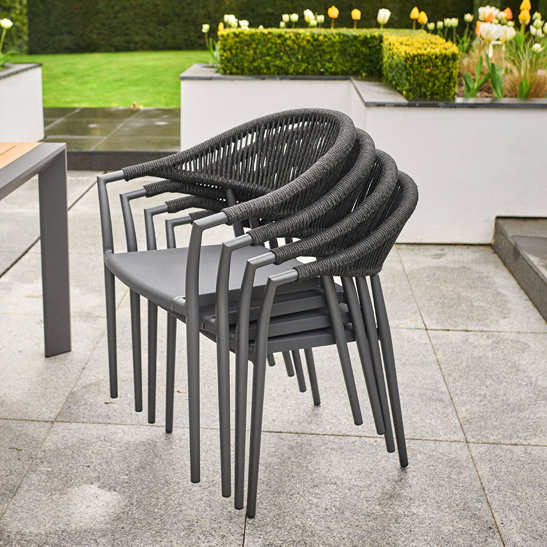Stacked gray patio chairs with woven backs rest on a stone-tiled patio, surrounded by a garden and flowerbeds in the background. The weather-proof aluminum frames and removable sun-proof cushions make them perfect additions to our Cloverly 8 Seat Rectangular Extending Dining Set with Ceramic Table in Charcoal.