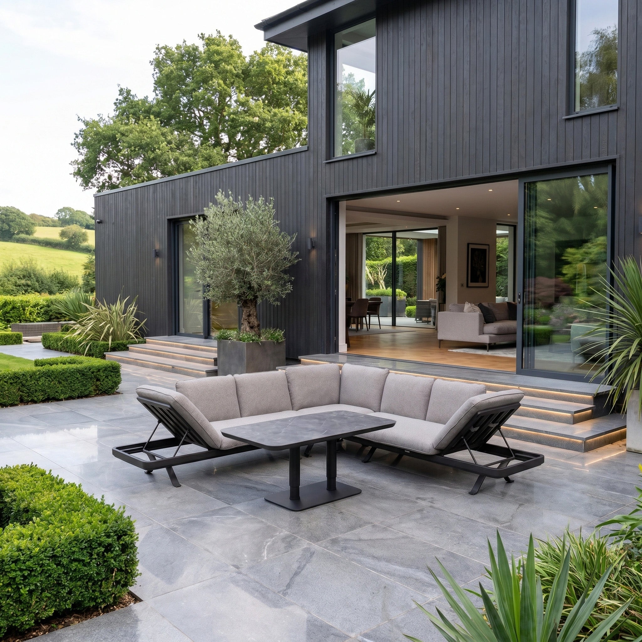 Modern patio with lush plants, black house, and large sliding glass doors, featuring outdoor luxury furniture like the Como Corner Sofa Set with Altura Rising Table in Truffle.