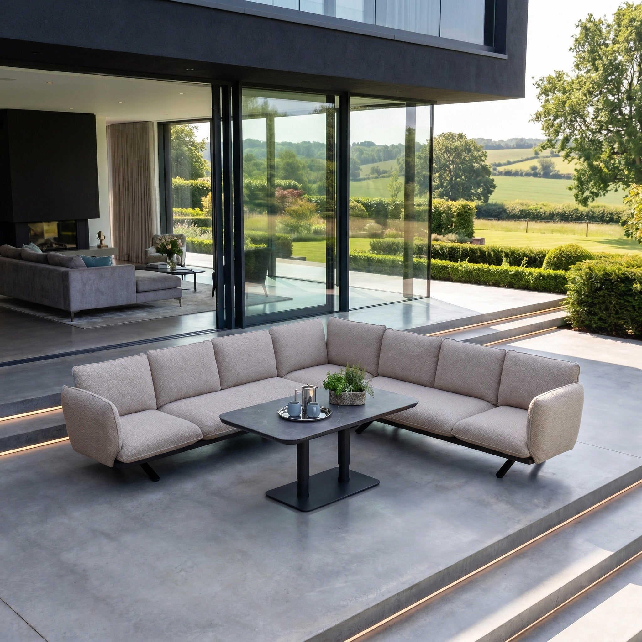 Modern patio showcasing the Como Corner Sofa Set with Altura Rising Table in Truffle—luxury outdoor furniture with a beige sectional, black rising table, and scenic countryside view in the background.