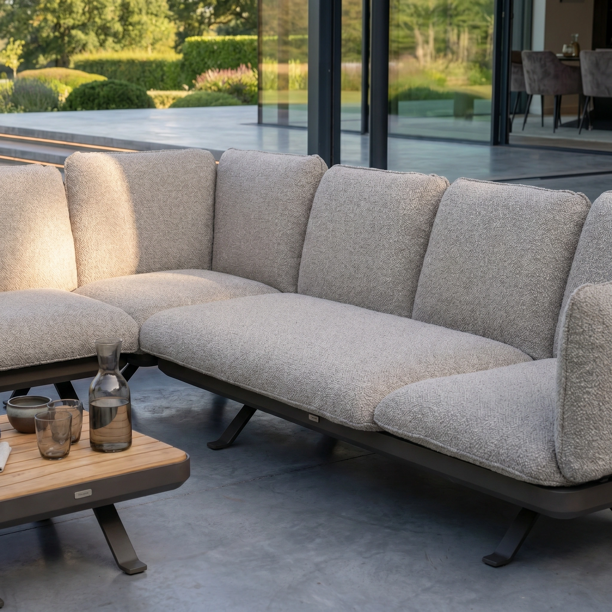 The Como Corner Sofa Set in Truffle brings elegance to your patio, nestled beside a glass-walled house and garden—an ideal luxury outdoor sofa for any stylish space.