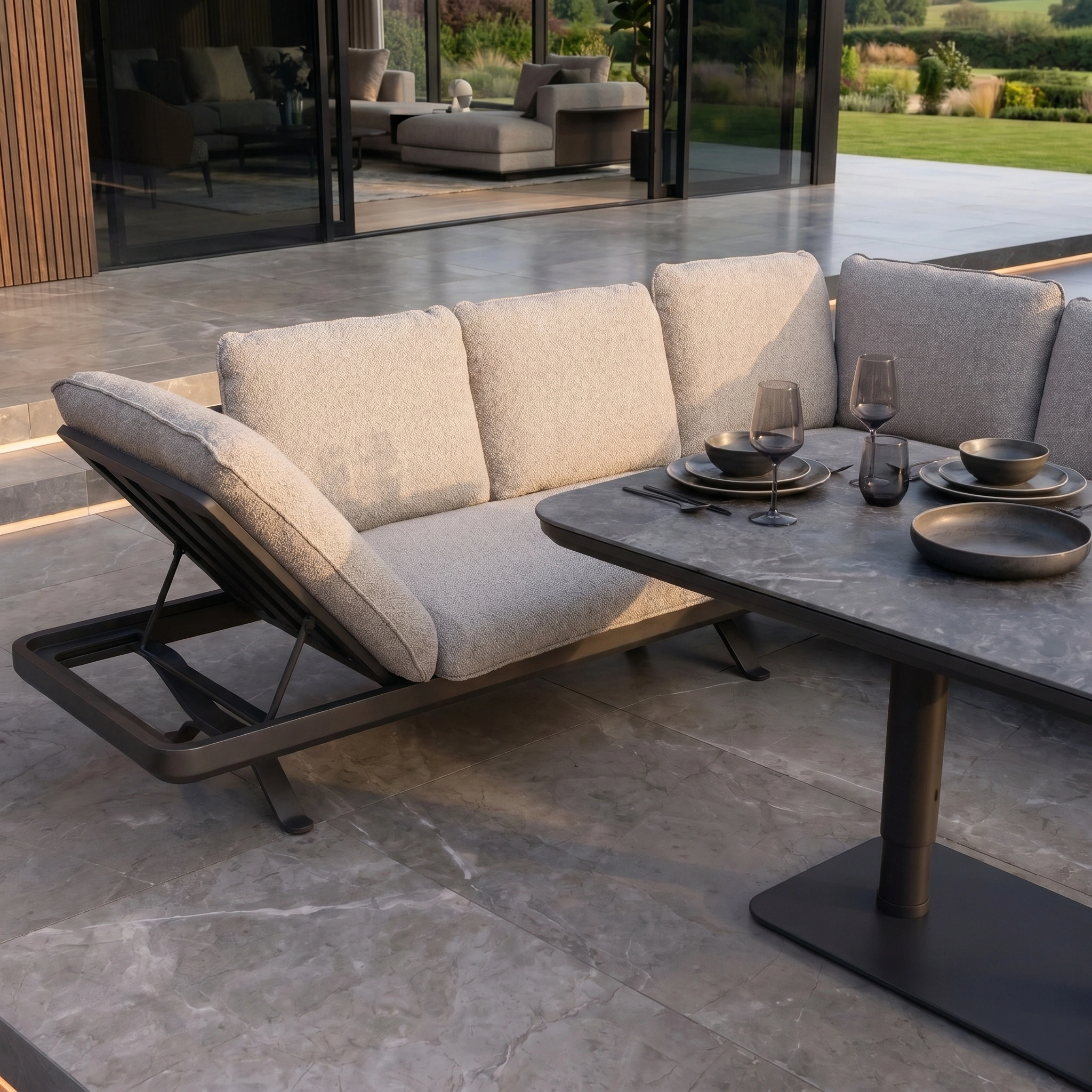 Modern outdoor patio featuring the Como Corner Sofa Set with Altura Rising Table in Truffle and tableware on tiled flooring near a garden—ideal for stylish, luxury outdoor living.