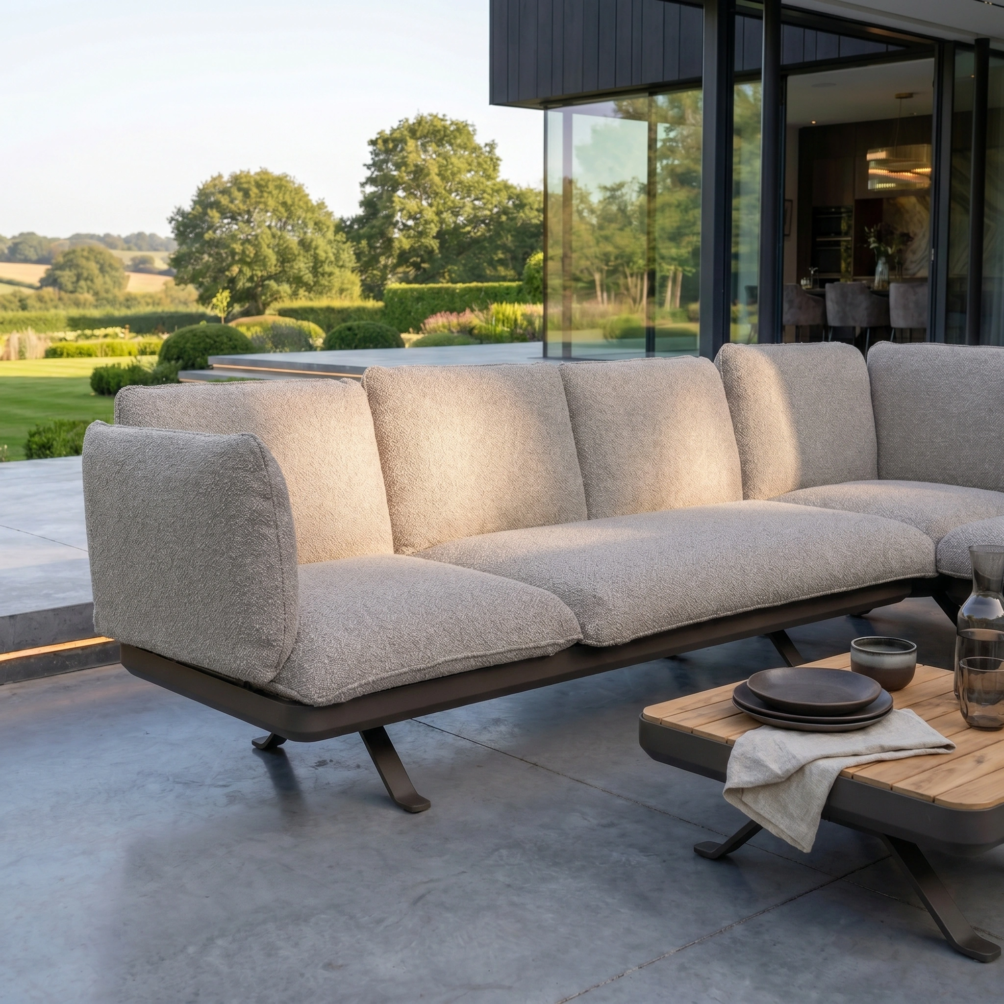 Modern outdoor patio featuring the Como Corner Sofa Set in Truffle, a luxurious light gray sofa with a wooden coffee table set on a concrete floor.