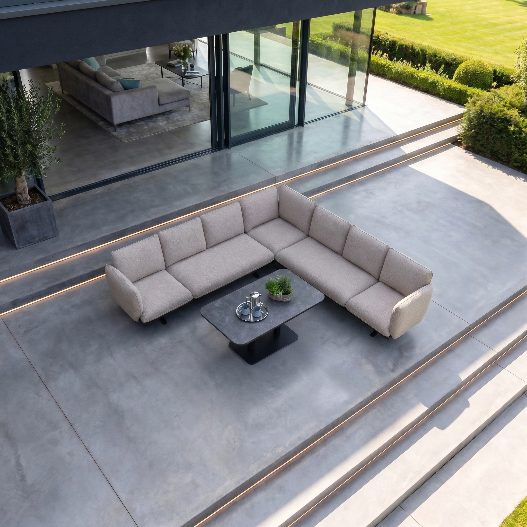 Modern outdoor patio with a beige Como Corner Sofa Set with Altura Rising Table in Truffle and a garden backdrop on concrete flooring—ideal for those seeking luxury outdoor furniture.