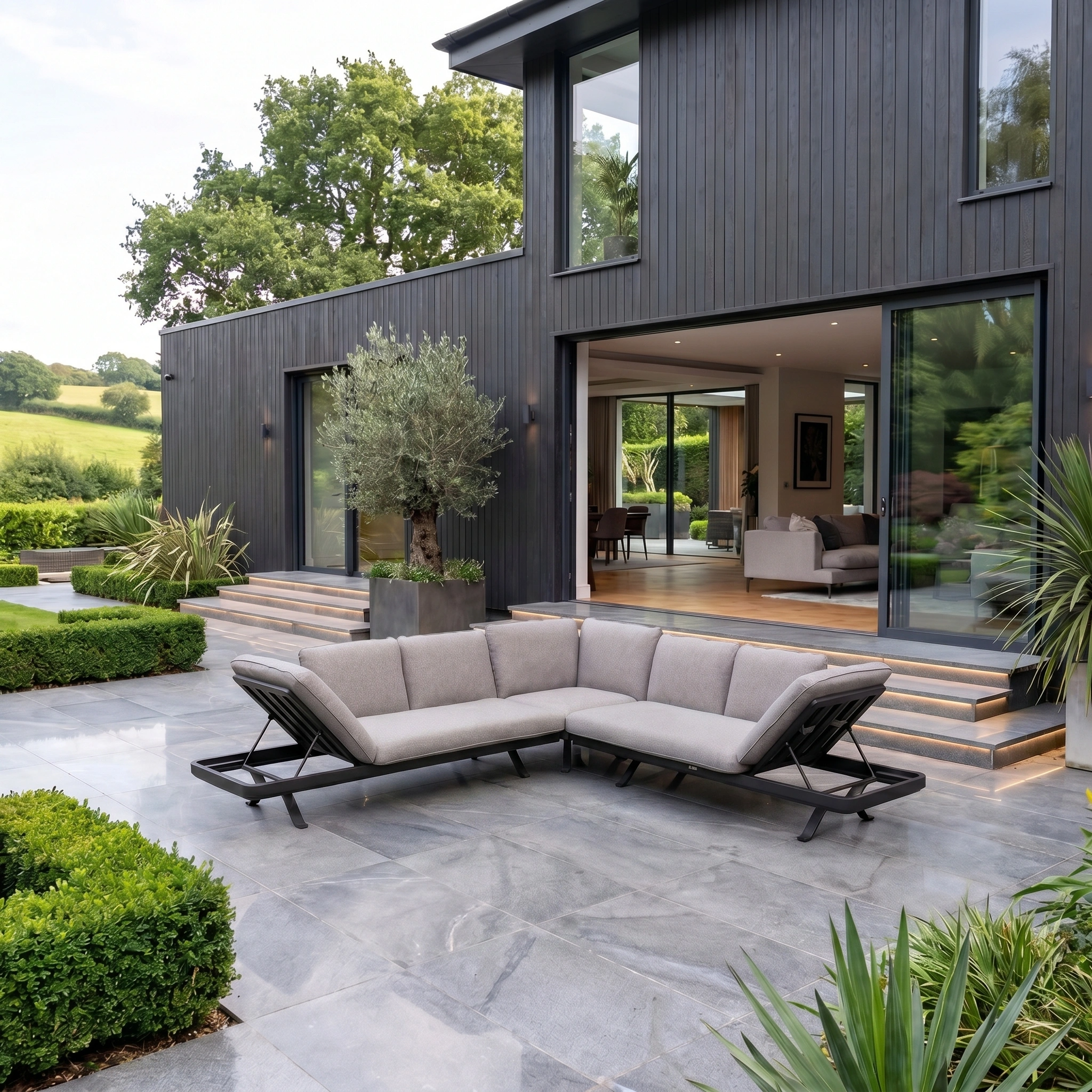 Modern gray house with large glass doors, patio seating featuring a Como Corner Sofa Set in Truffle, and landscaped greenery on a sunny day.