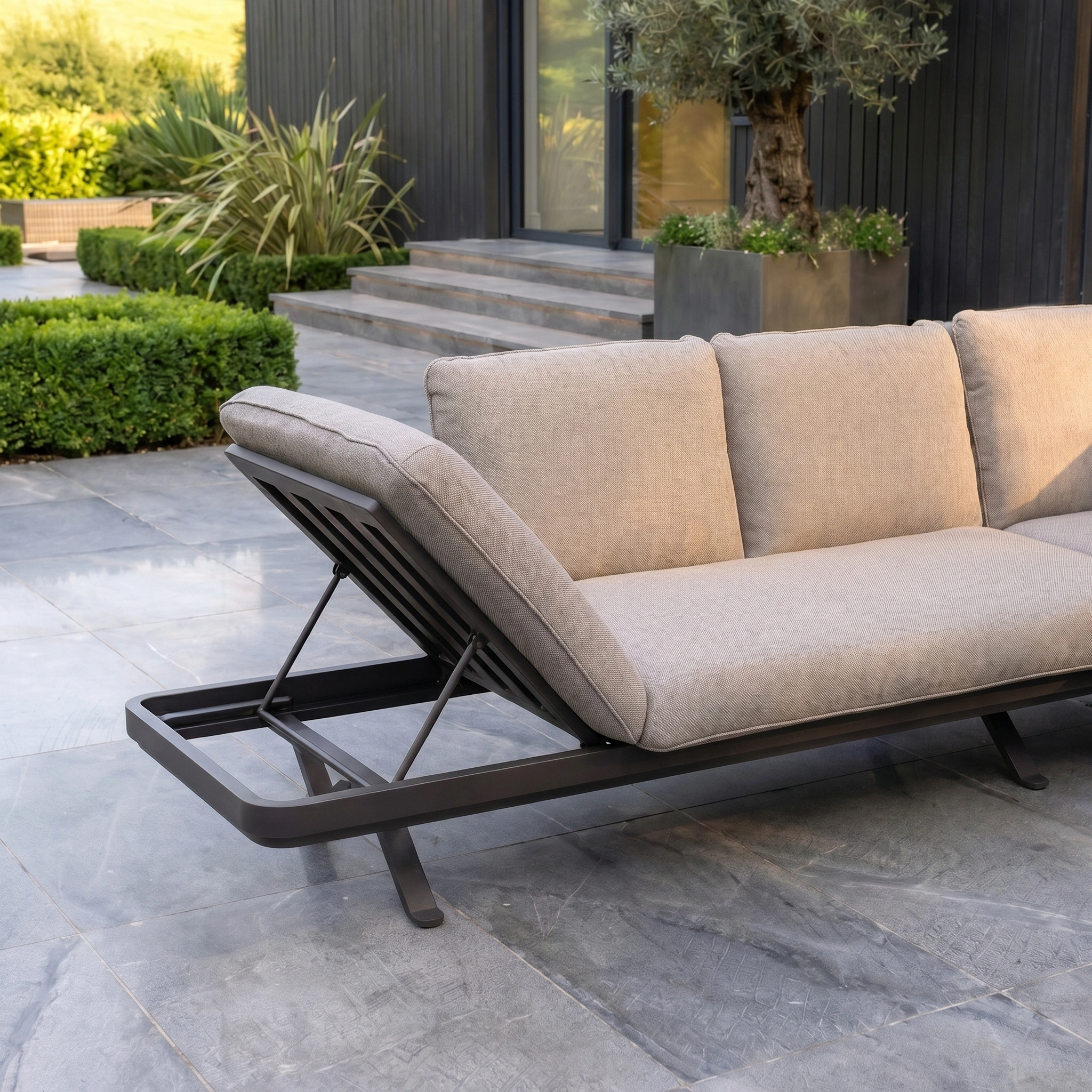 The Como Corner Sofa Set in Truffle with a black metal frame sits on a stone patio, paired with a teak coffee table, in front of a contemporary house.