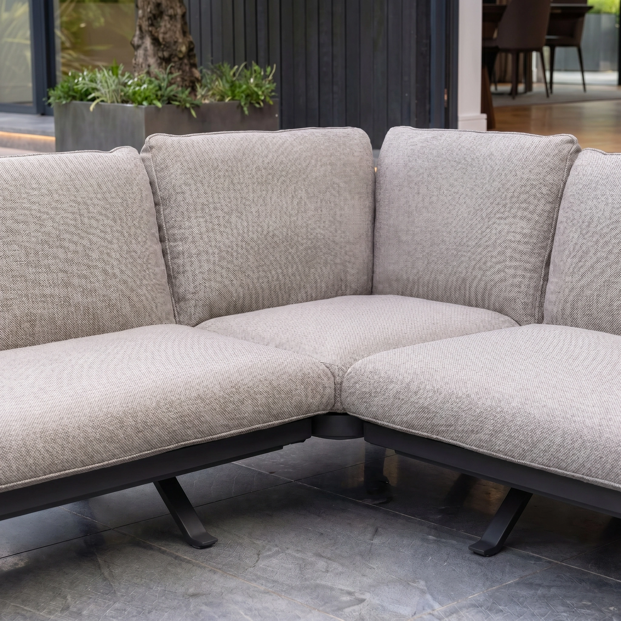 The Como Corner Sofa Set in Truffle features thick cushions on a patio and pairs with a teak coffee table. Surrounded by plants and a welcoming indoor space, this outdoor luxury sofa set adds elegance to any gathering.