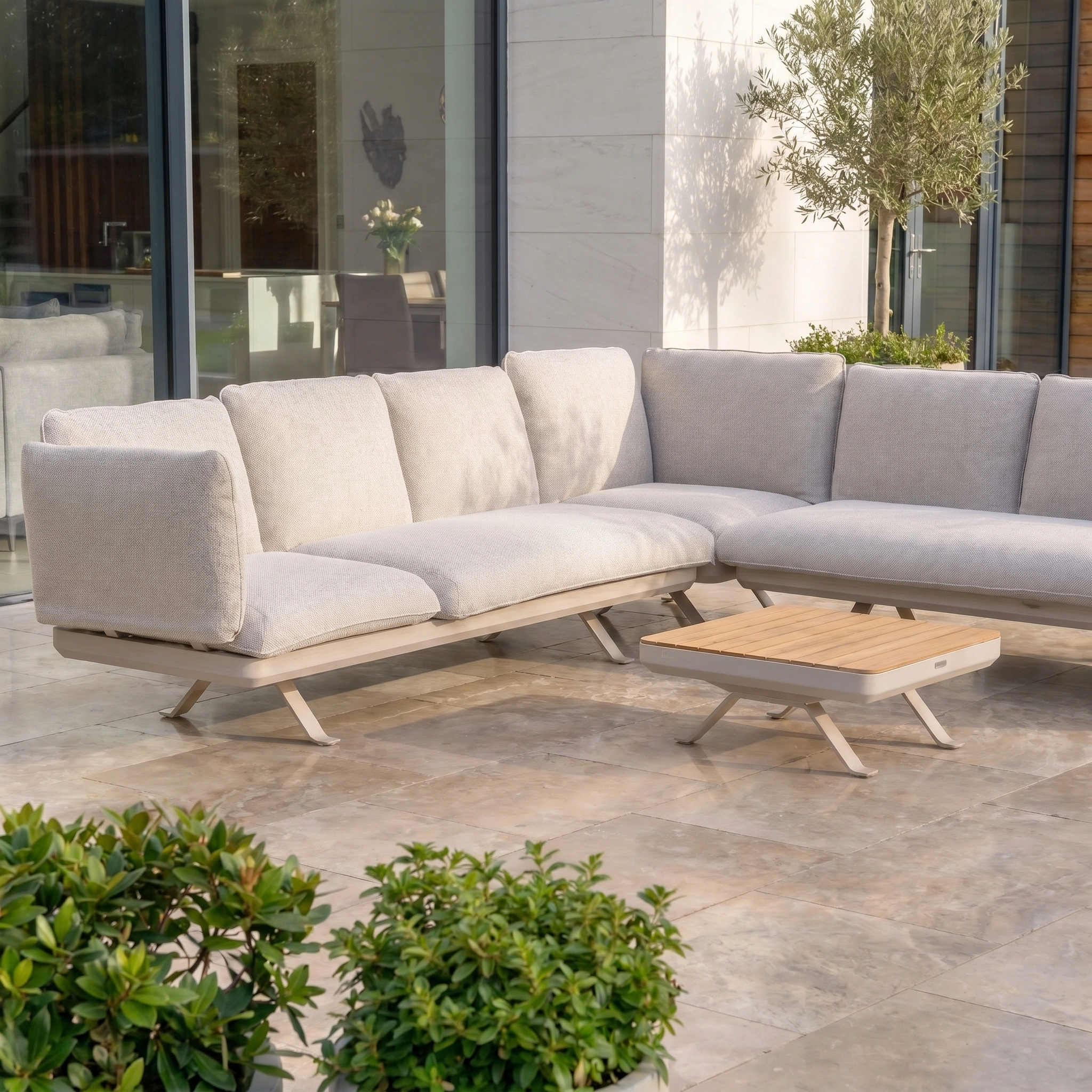 Modern outdoor patio with a Como Corner Sofa Set in Oatmeal, a wooden coffee table, and green plants on stone tiles—stylish modular outdoor furniture ideal for a relaxing retreat.