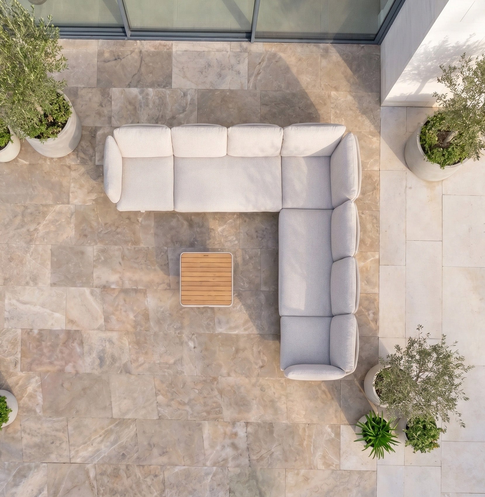 The Como Corner Sofa Set in Oatmeal sits on a stone patio with a wooden table and potted plants, creating an inviting outdoor luxury space.
