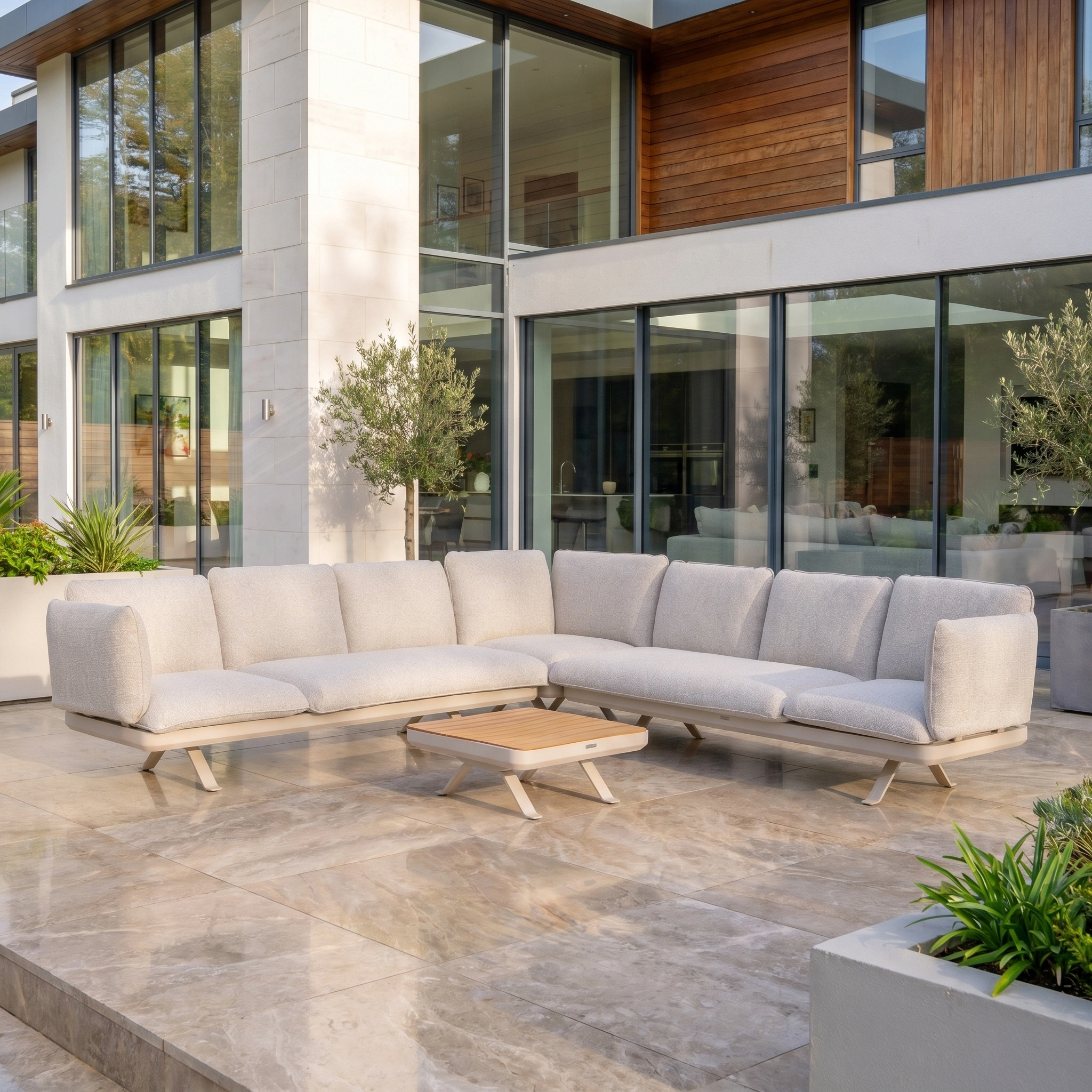 Modern outdoor patio with the Como Corner Sofa Set in Oatmeal, a beige modular design, and a low wooden table in front of a glass-windowed house.