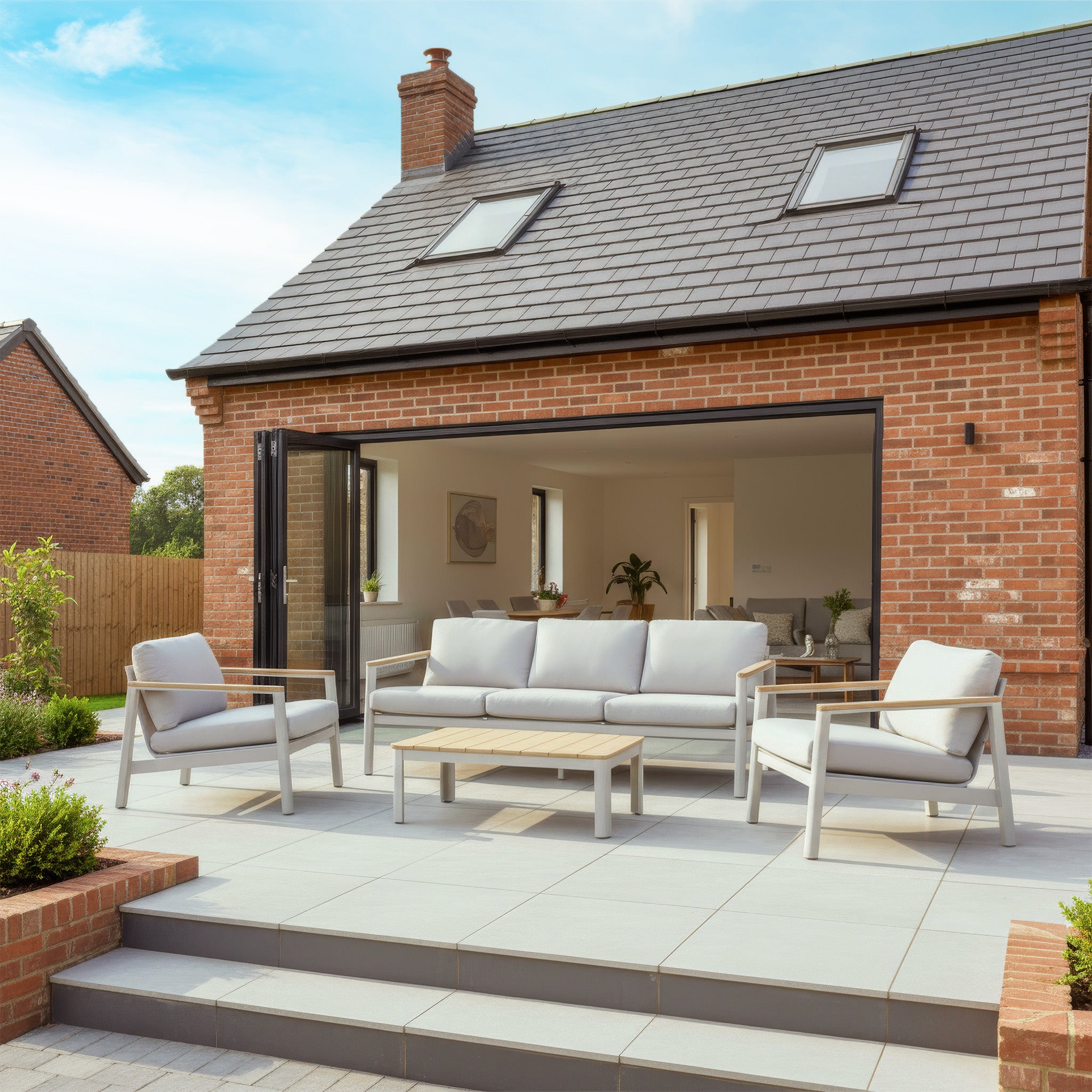 Modern patio with the weather-resistant Cora 3 Seat Sofa Set in Latte in front of a brick house, featuring large glass doors and a tiled roof.