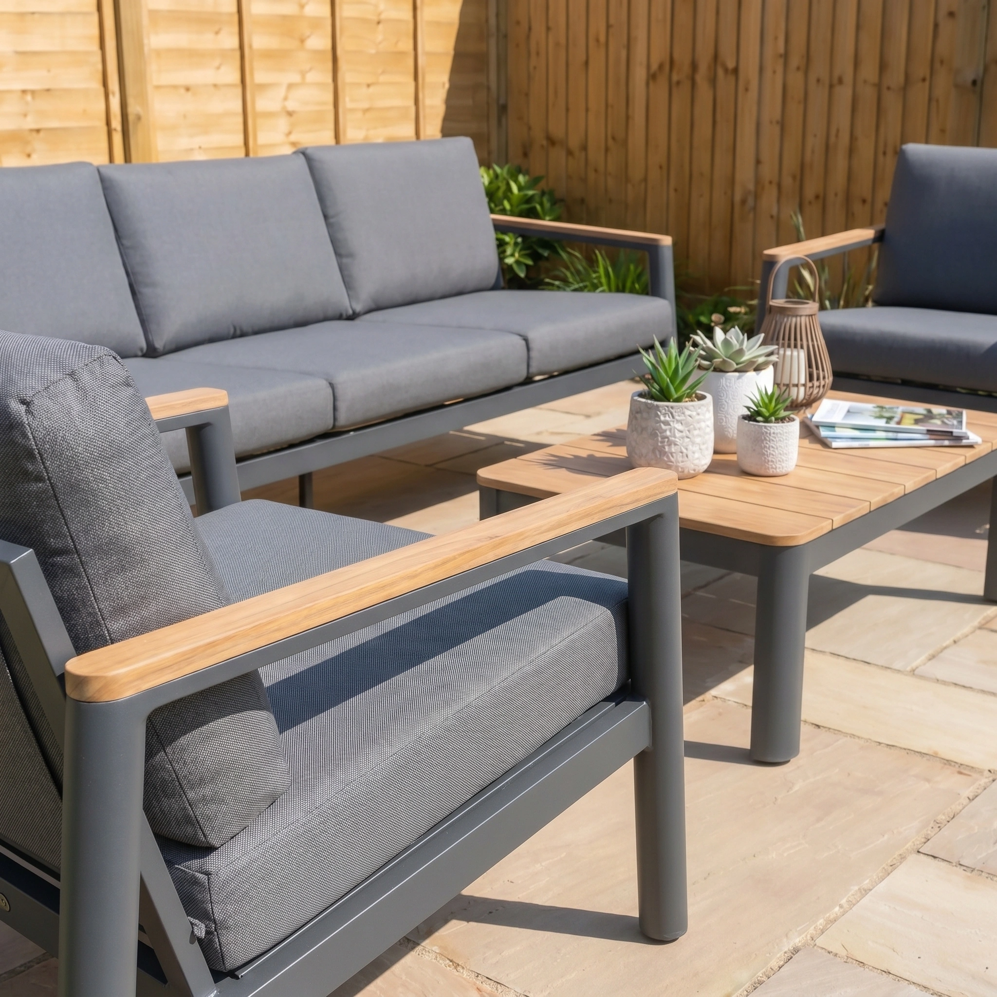 The Cora 3 Seat Sofa Set in Grey features wooden arms, a coffee table, potted plants, and magazines on a stone patio—stylish and weather resistant garden furniture ideal for any modern outdoor space.