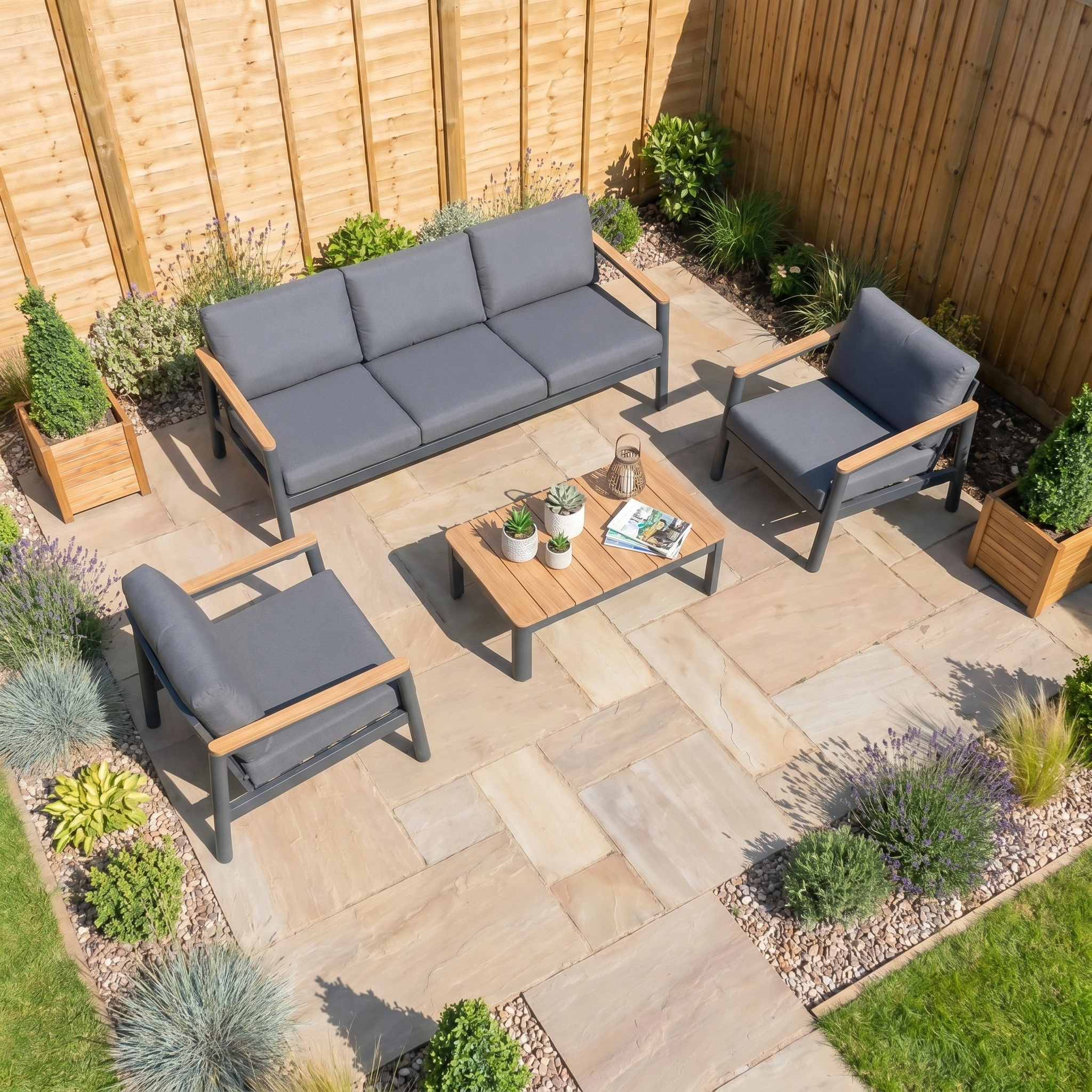 Modern patio with the Cora 3 Seat Sofa Set in Grey and coffee table on stone tiles, surrounded by plants and a wooden fence. This weather-resistant set offers style and durability for an inviting outdoor space.