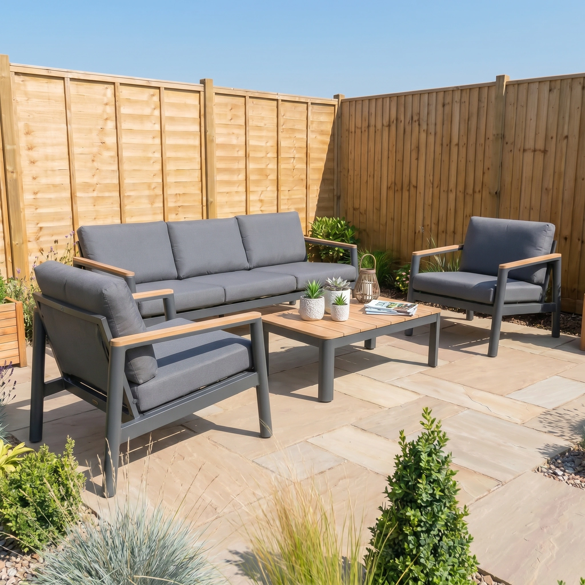 Patio featuring the Cora 3 Seat Sofa Set in Grey—an elegant, weather-resistant outdoor furniture set with sofa, chairs, and coffee table—surrounded by lush plants and wooden fencing.