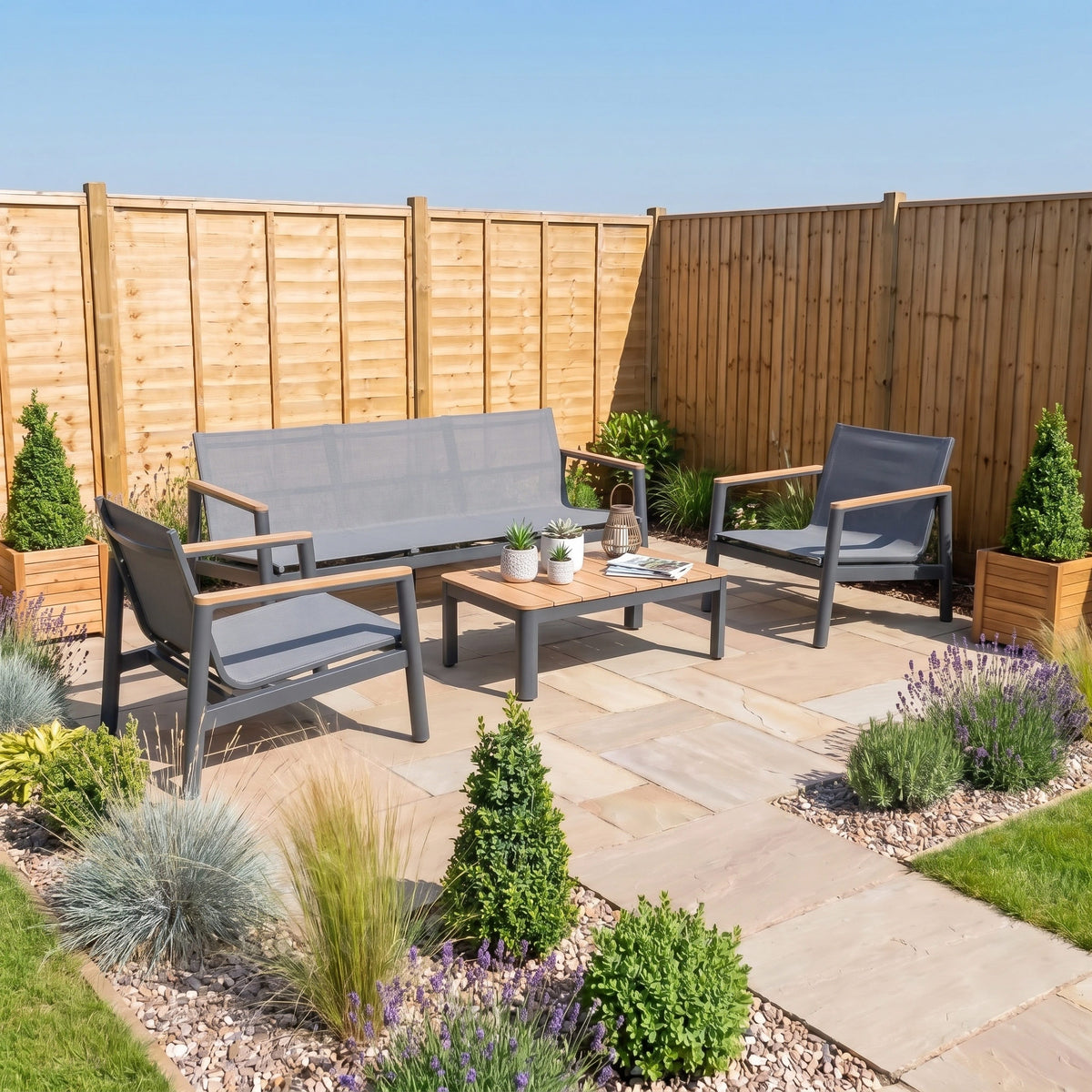 Patio with weather resistant outdoor furniture, featuring the modern Cora 3 Seat Sofa Set in Grey, a wooden coffee table, and potted plants on stone tiles in a fenced backyard.