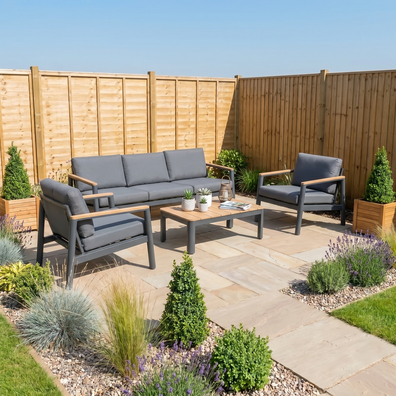 The Cora 3 Seat Sofa Set in Grey features weather-resistant cushions and is shown on a stone patio with lush plants and wooden fencing.