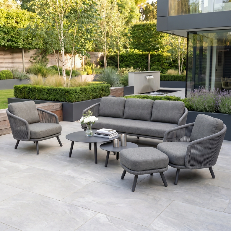 Create a stylish outdoor space with the Dekota 3 Seat Rope Sofa Set in Charcoal, featuring grey olefin rope weave seating, powder-coated aluminium frame, coffee tables, footstool, and lush garden greenery.